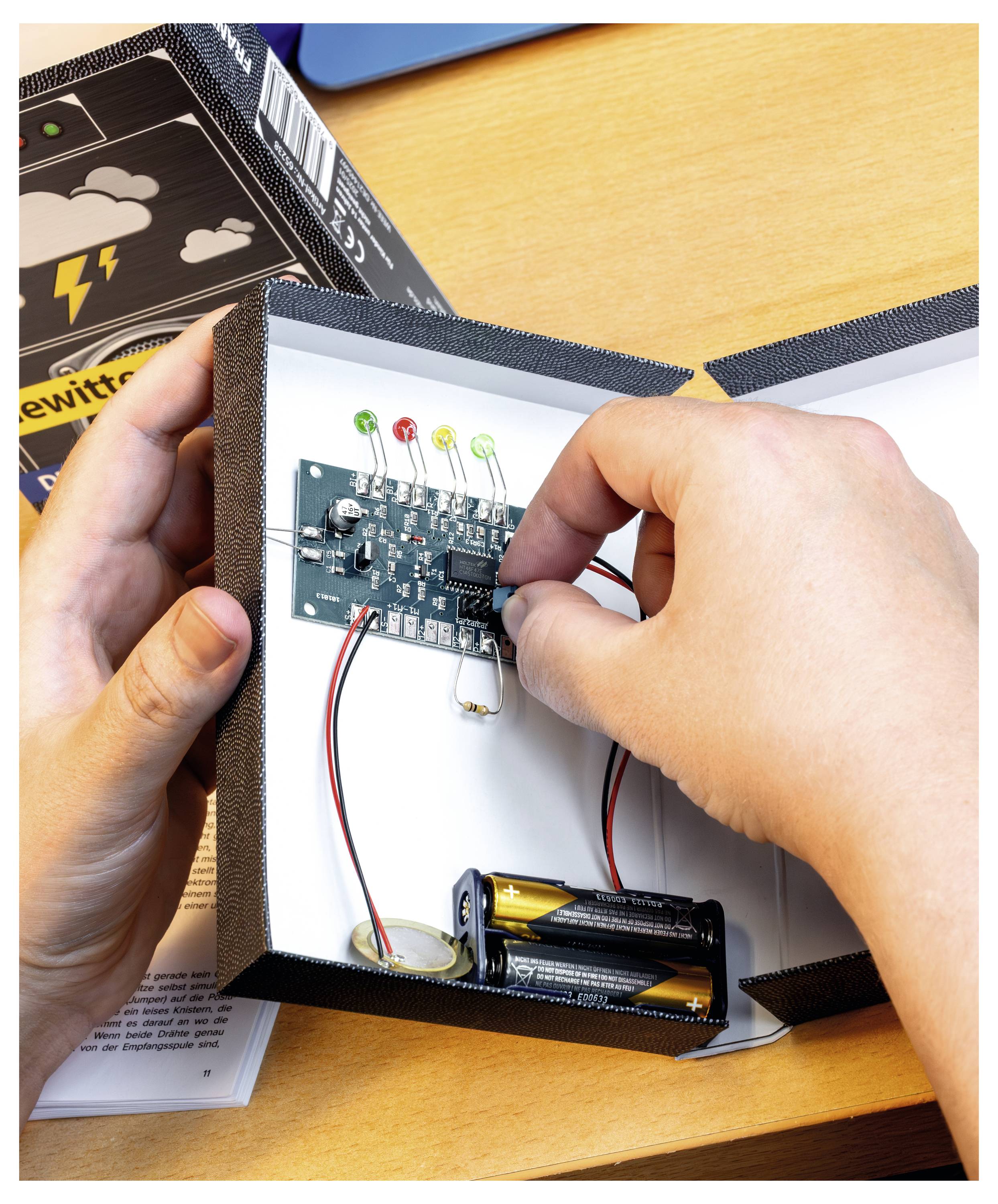Hände passen eine Leiterplatte in einer Kiste an; Batterie und LEDs sind sichtbar. Ein Buch mit dem Titel 