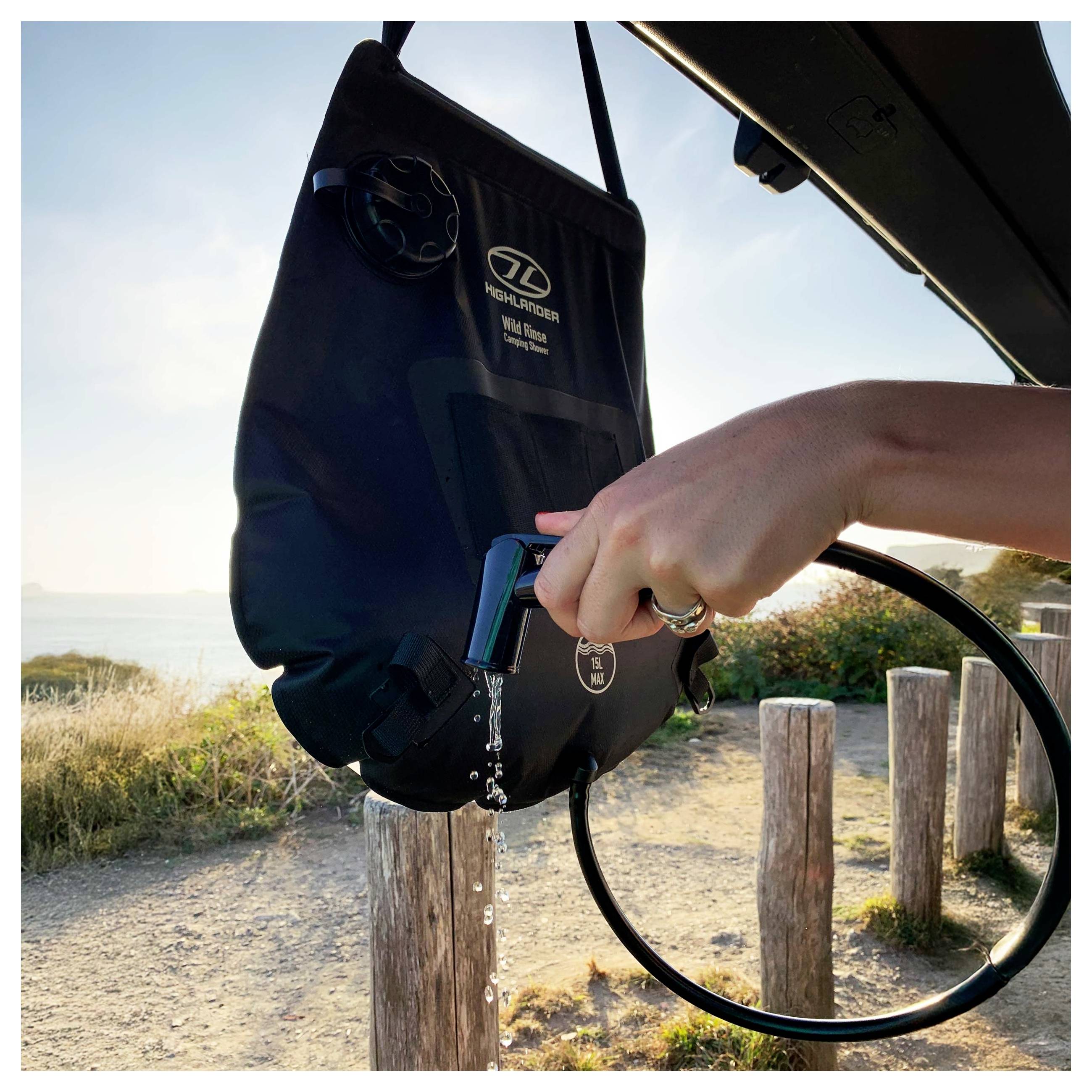 Eine Person nutzt einen tragbaren Wasserbeutel, um Wasser auf einen Küstenweg zu gießen, der von Gras und Holzpfosten umgeben ist.