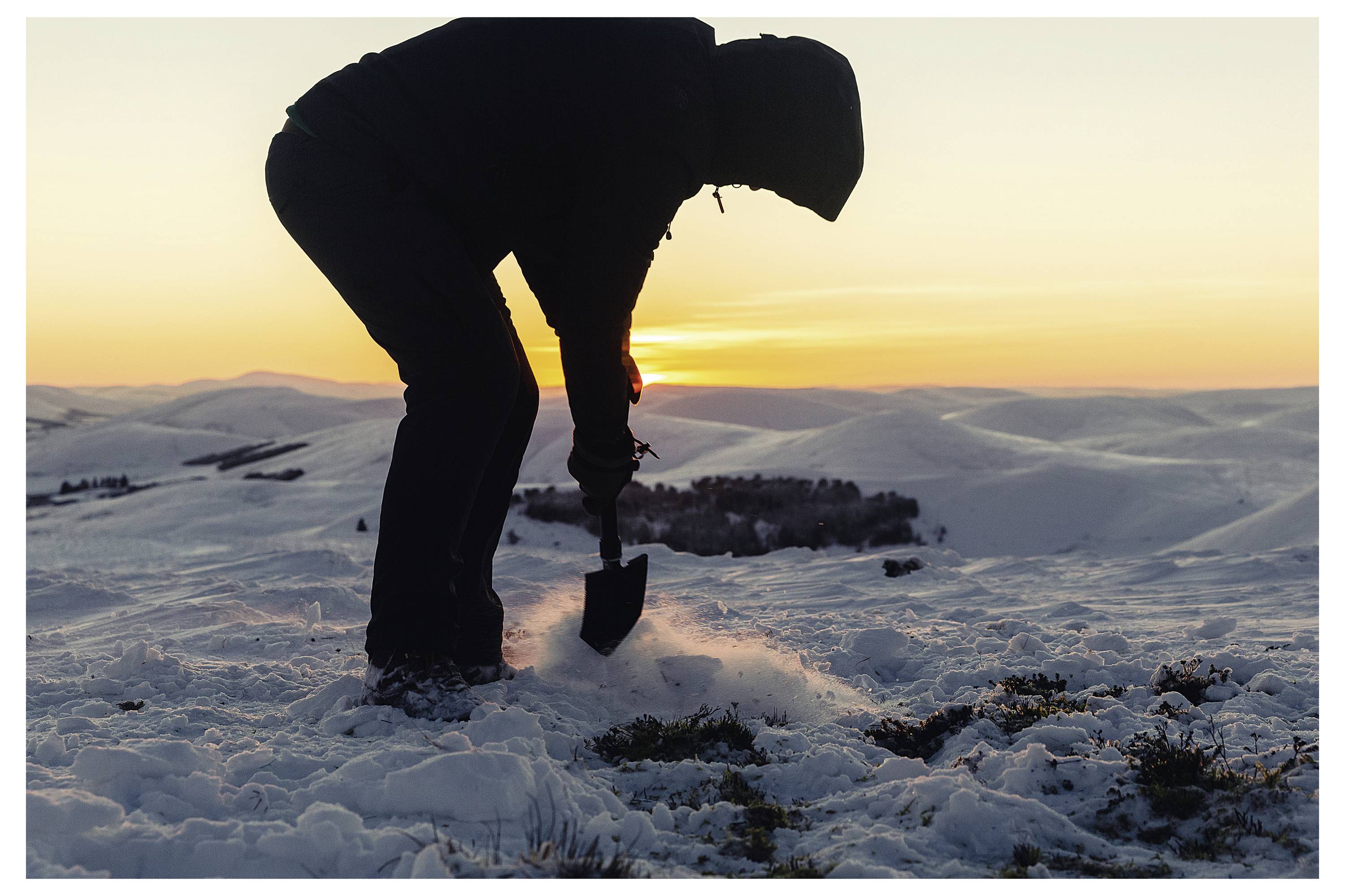 Eine Person in Winterkleidung schaufelt Schnee bei Sonnenuntergang in einer schneebedeckten Landschaft und betont damit Einsamkeit und die Weite der Umgebung.