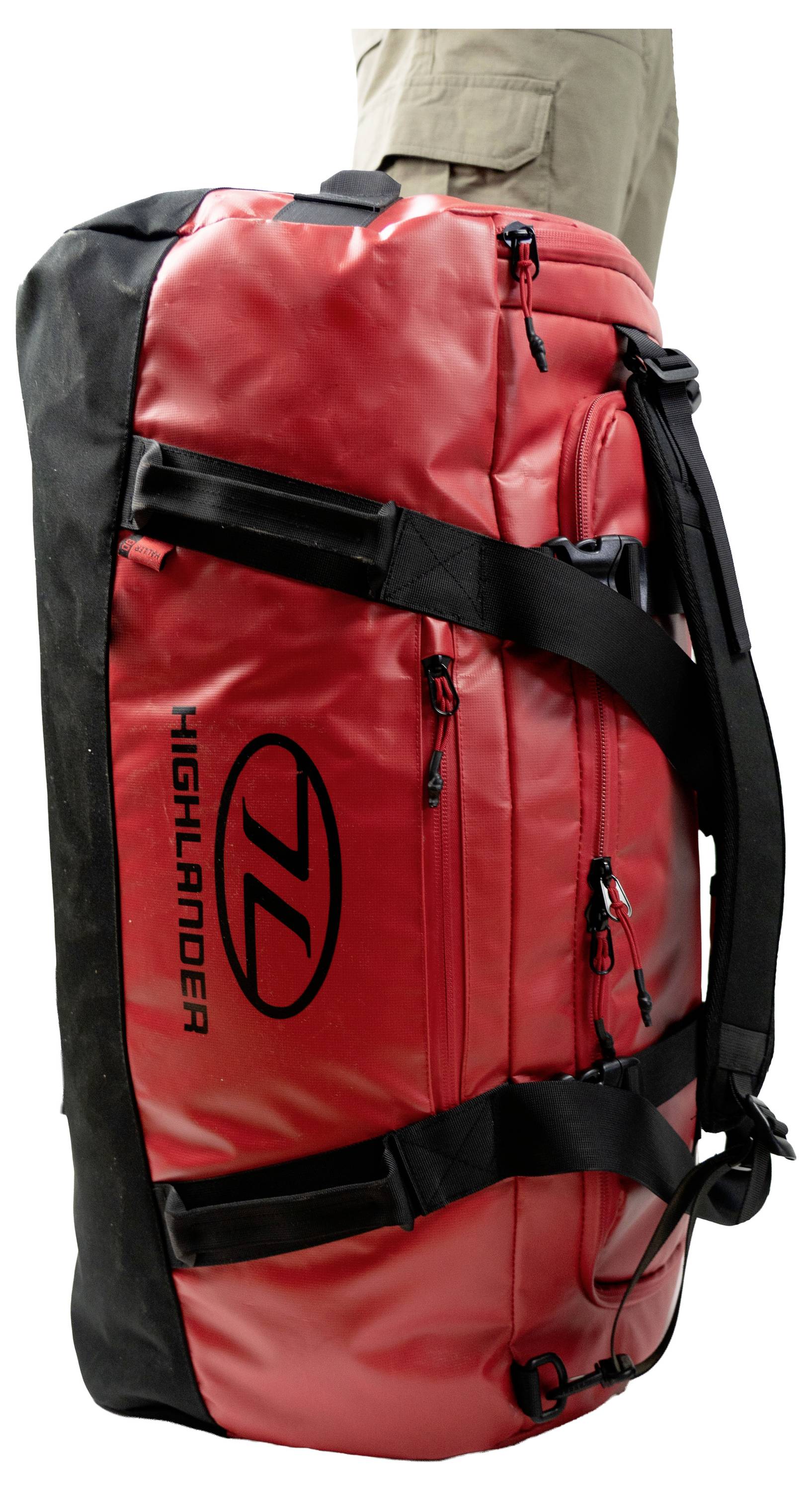 Ein großer, rot und schwarz karierter Highlander-Sporttasche, der aufrecht steht, mit mehreren Riemen und Reißverschlüssen, neben einer Person in beigen Hosen.