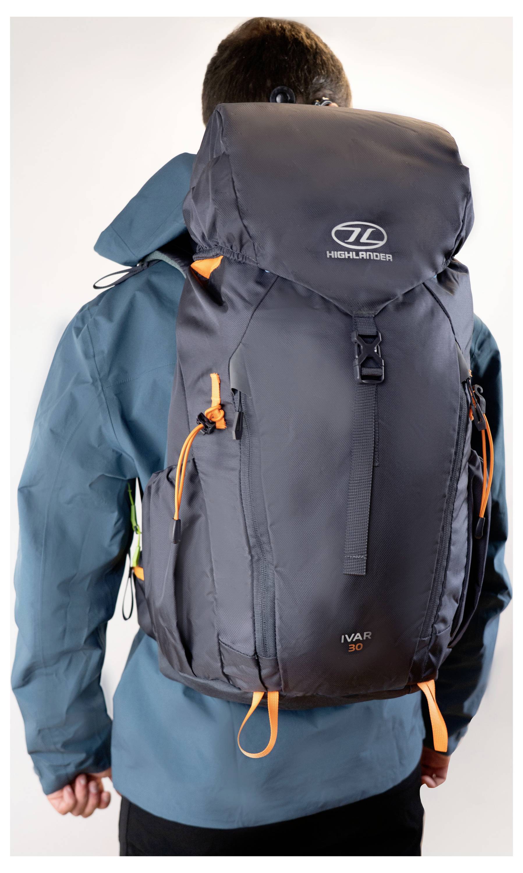 Eine Person in einer blauen Jacke wird von hinten gesehen und trägt einen großen, dunkelgrauen Wanderrucksack mit orangefarbenen Riemen.