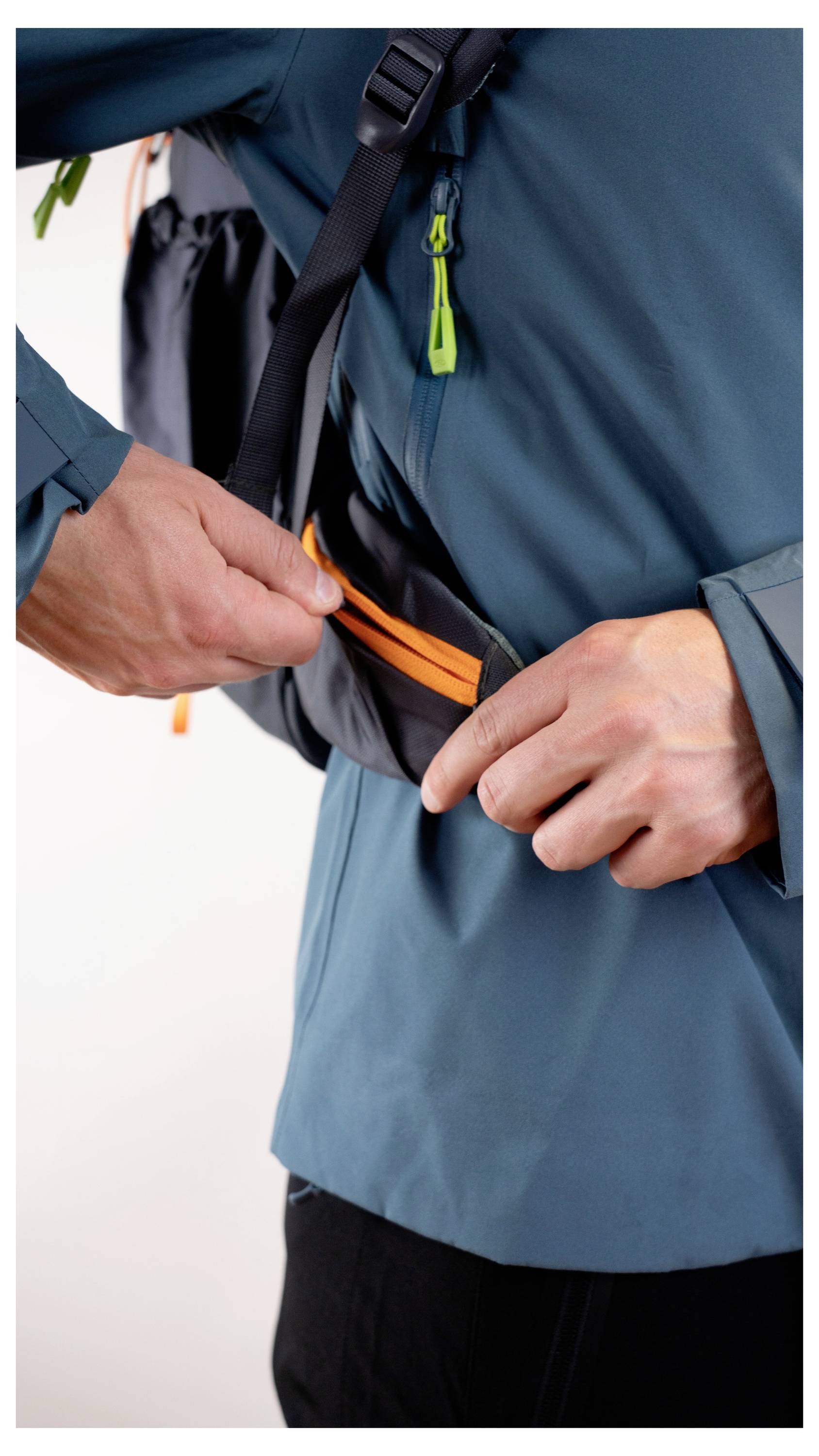 Eine Person in einer blaugrünen Jacke reißt einen kleinen orangefarbenen Reißverschlusstasche an einem Rucksackträger zu.