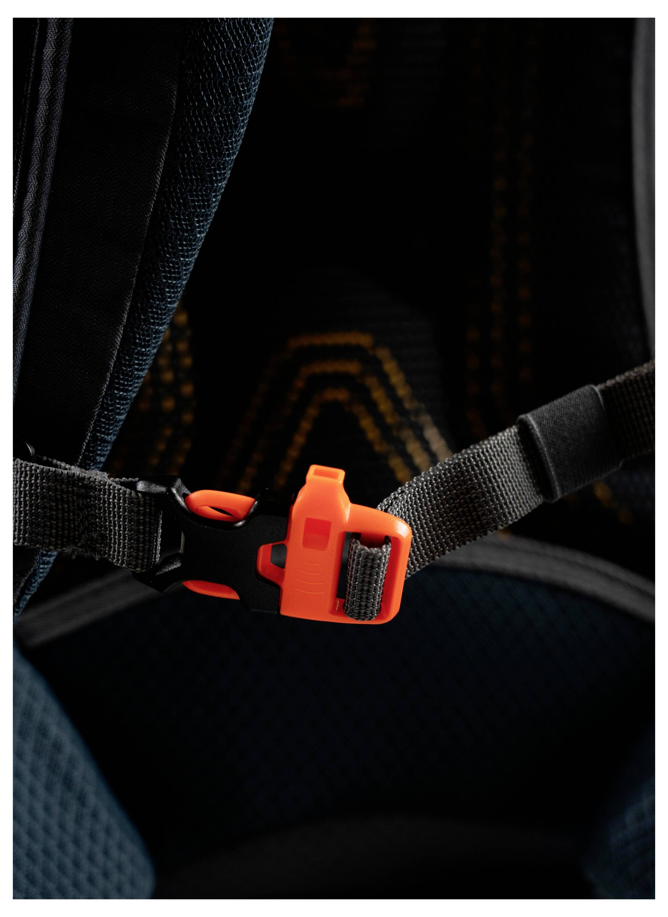 Eine orange-schwarze Kunststoffschnalle befestigt einen schwarzen Riemen auf einem dunklen Stoffhintergrund, wahrscheinlich Teil eines Rucksacks oder Geschirrs.