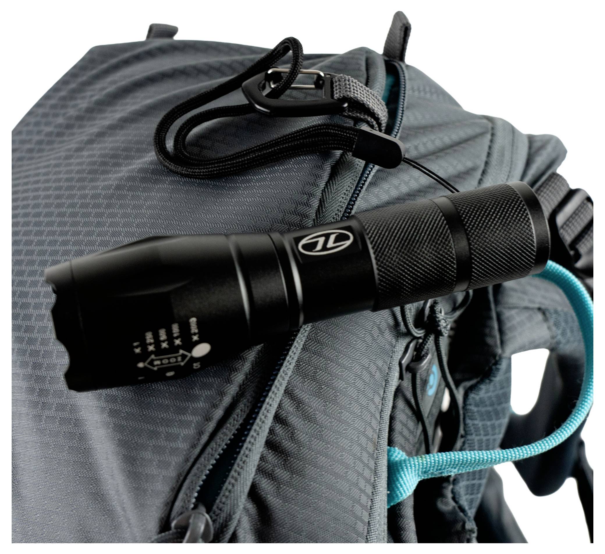 Eine schwarze Taschenlampe, die an dem Riemen eines blaugrauen Rucksacks befestigt ist und die wichtigsten Ausrüstungsgegenstände für Outdoor-Aktivitäten hervorhebt.