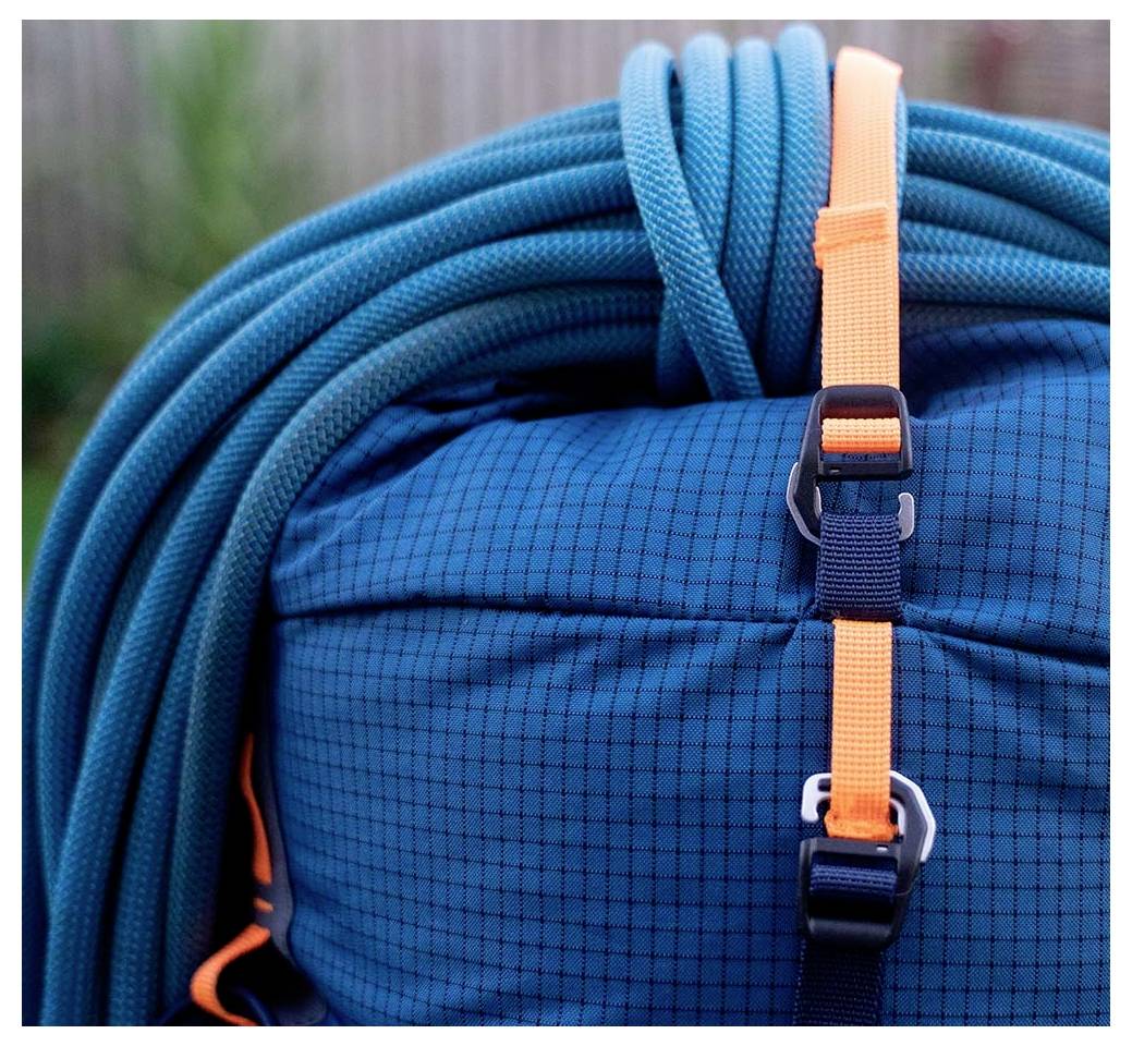 Ein blauer Kletterrucksack mit angebrachtem aufgerolltem Seil und orangefarbenen Riemen. Der Hintergrund ist verschwommen und deutet auf eine Außenumgebung hin.