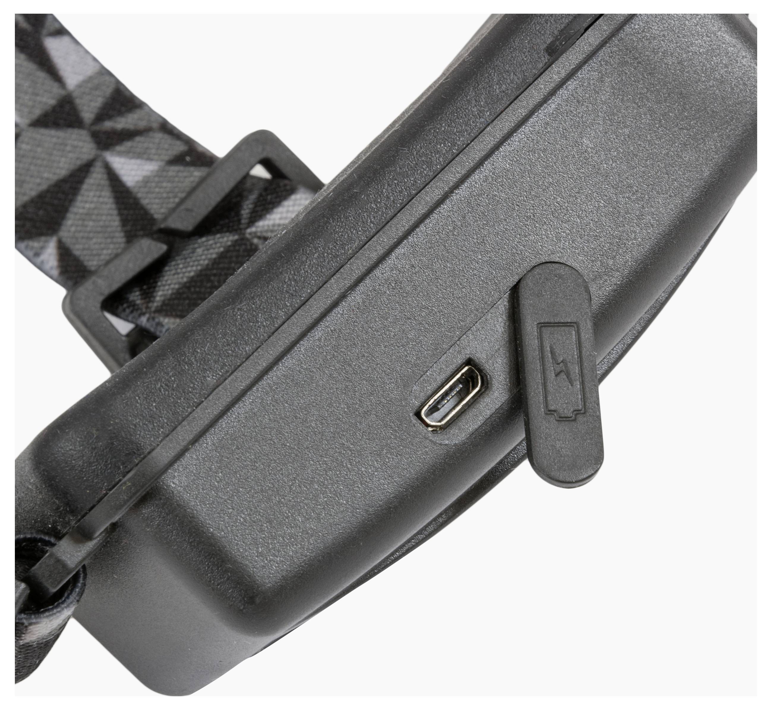 Eine Nahaufnahme eines Ladeanschlusses auf einem schwarzen elektronischen Gerät mit einem gemusterten Riemen und einer angebrachten Gummiabdeckung mit einem Ladesymbol.