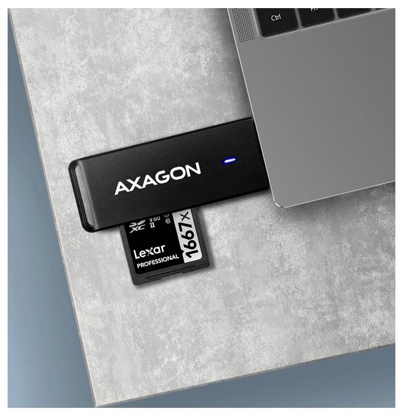 AXAGON USB-Kartenleser in einen Laptop eingesteckt, mit einer eingelegten Lexar Professional 1667x Speicherkarte auf einer grauen Oberfläche.
