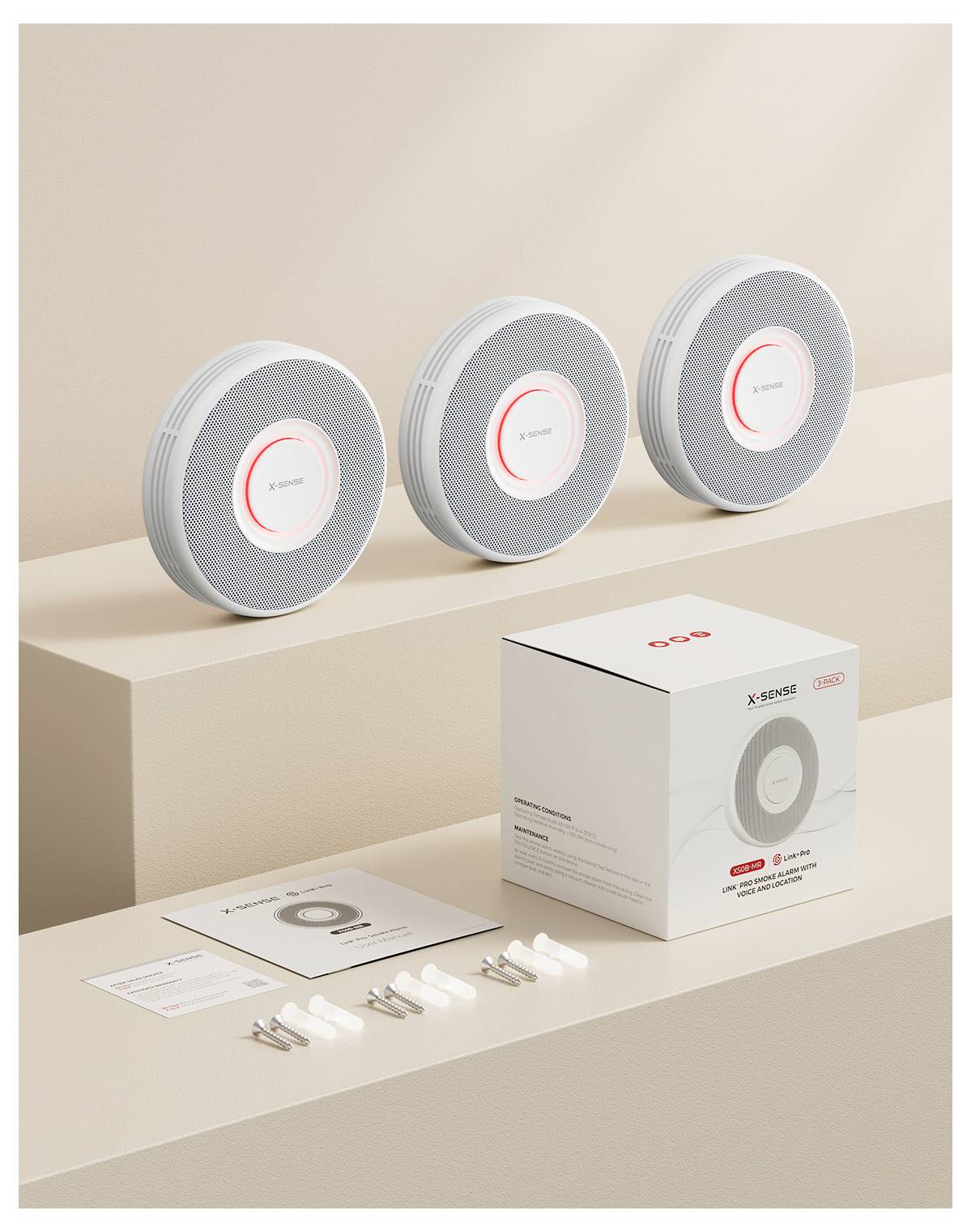 Drei kreisförmige Rauchmelder sind in einer Reihe auf einer beigen Oberfläche angeordnet. Davor befindet sich ein boxed Set mit Handbüchern und Montagezubehör.