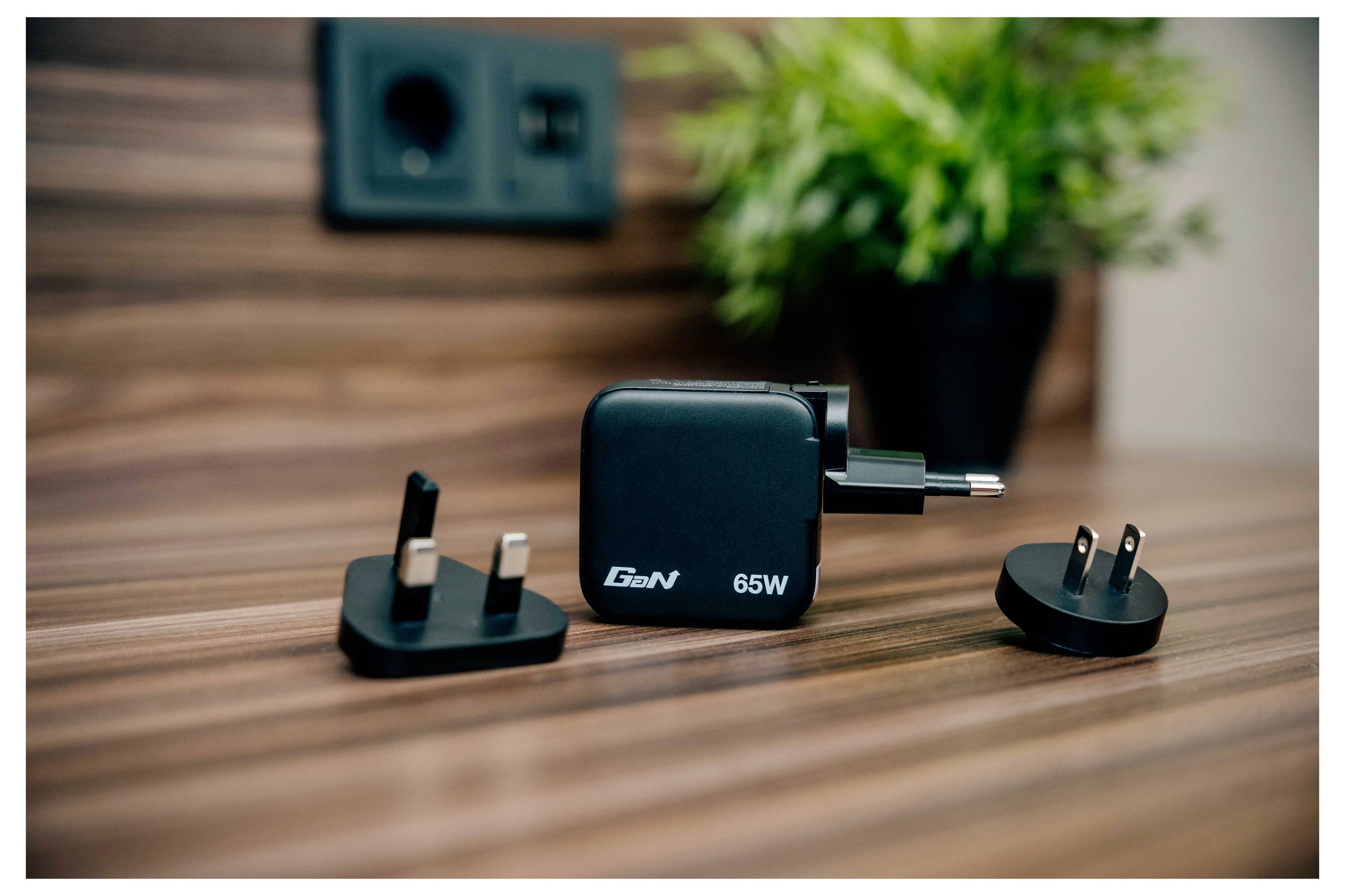 Ein schwarzes GaN-Ladegerät mit 65 W und drei austauschbaren Steckeradaptern sitzt auf einer Holzoberfläche. Eine Steckdose und eine Topfpflanze sind im Hintergrund.