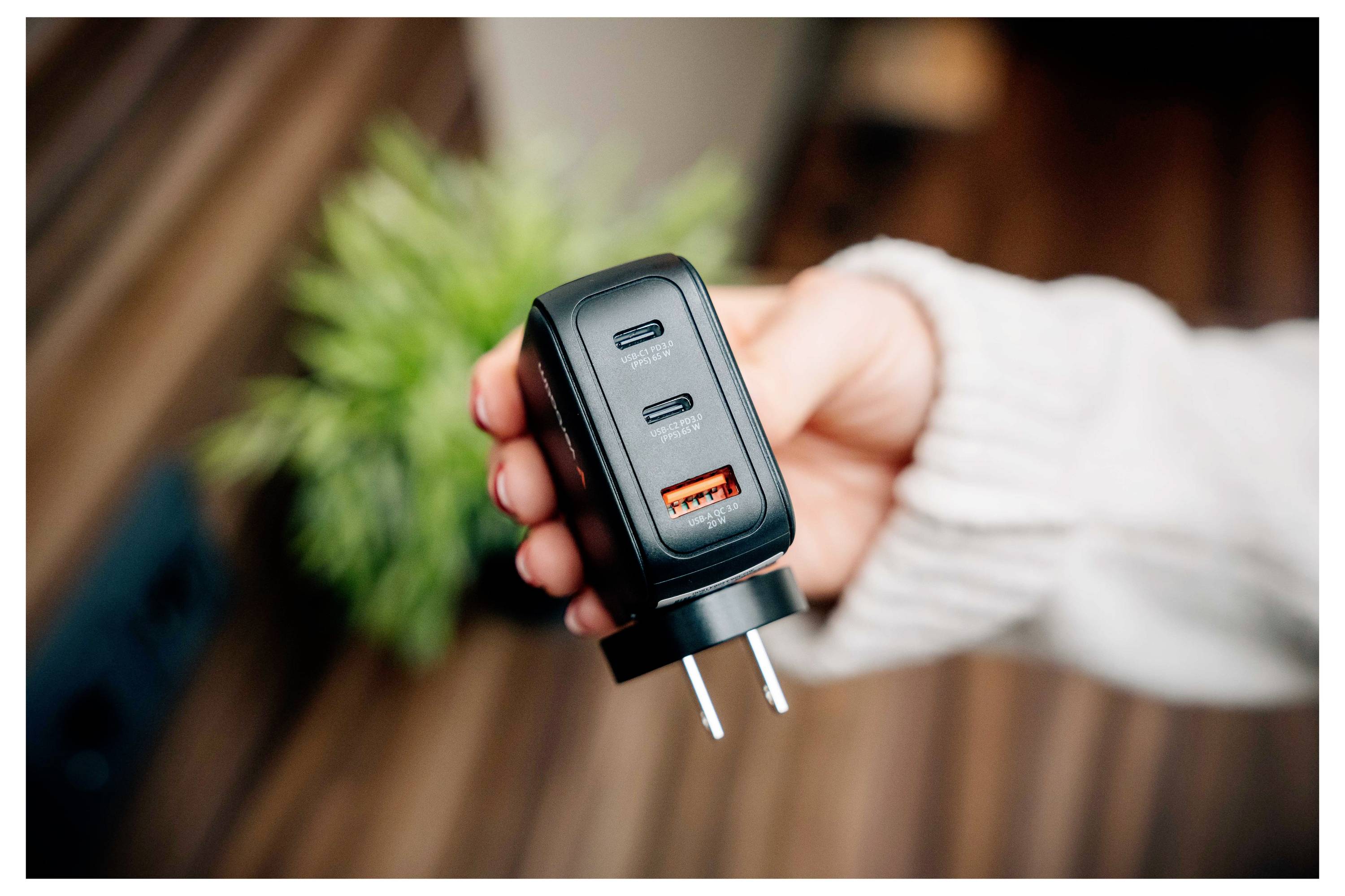 Eine Hand hält ein schwarzes Wandladegerät mit zwei USB-C-Anschlüssen und einem USB-A-Anschluss, mit einer unscharfen grünen Pflanze und einer Holzoberfläche im Hintergrund.