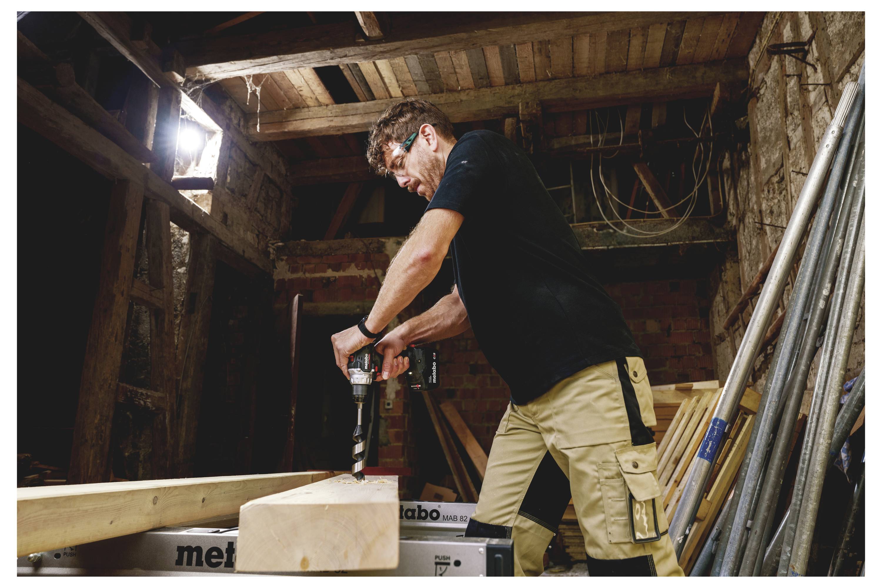 Ein Mann, der einen Elektroschrauber benutzt, um ein Loch in einen Holzblock in einer Werkstatt oder einem Bauschuppen zu bohren, mit Werkzeugen und Holz um ihn herum.
