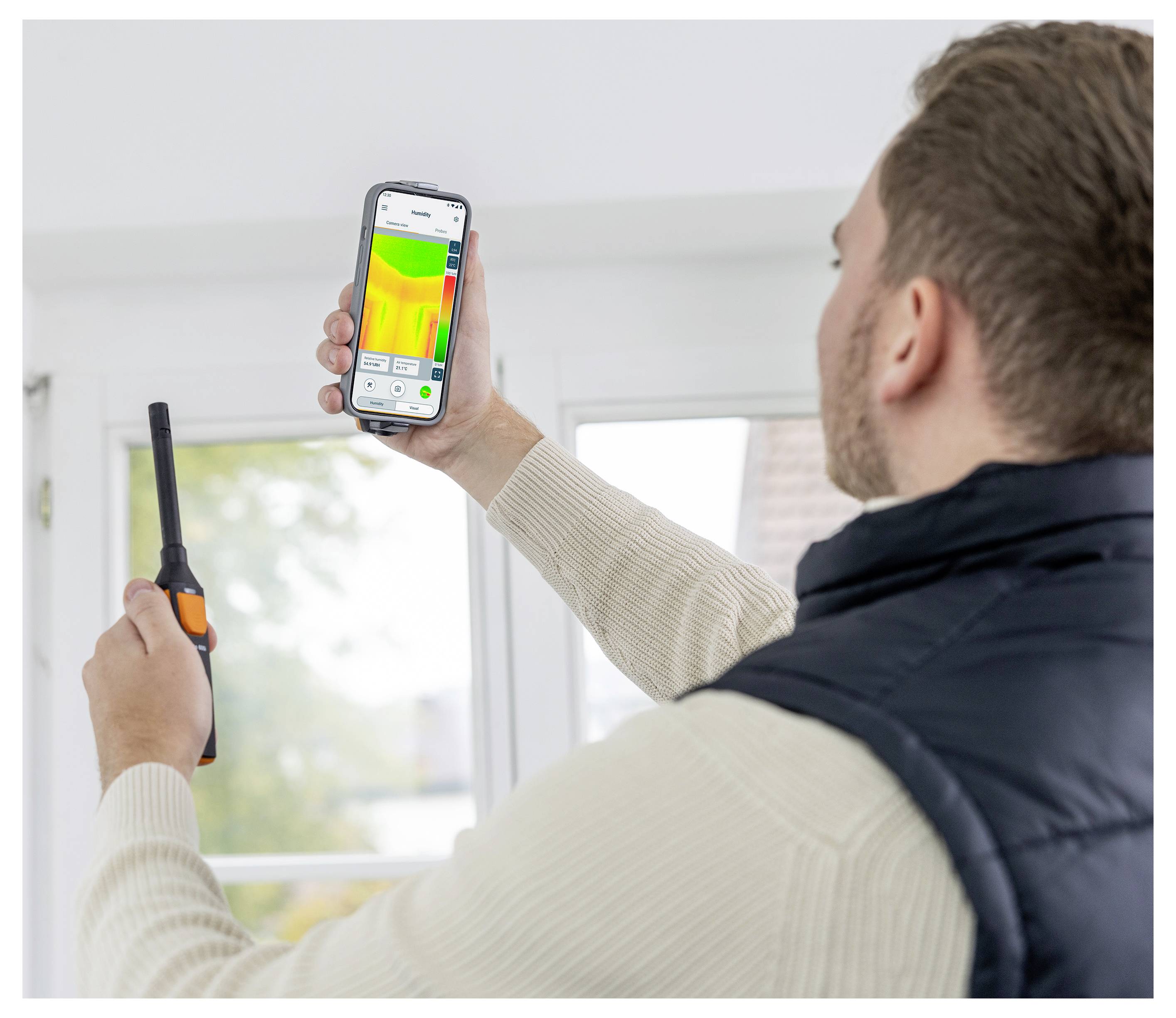 Eine Person hält ein Smartphone mit einer Wärmebildkamera-App in der Hand, während sie ein tragbares Gerät benutzt. Sie scheint drinnen nach Temperaturschwankungen zu suchen.