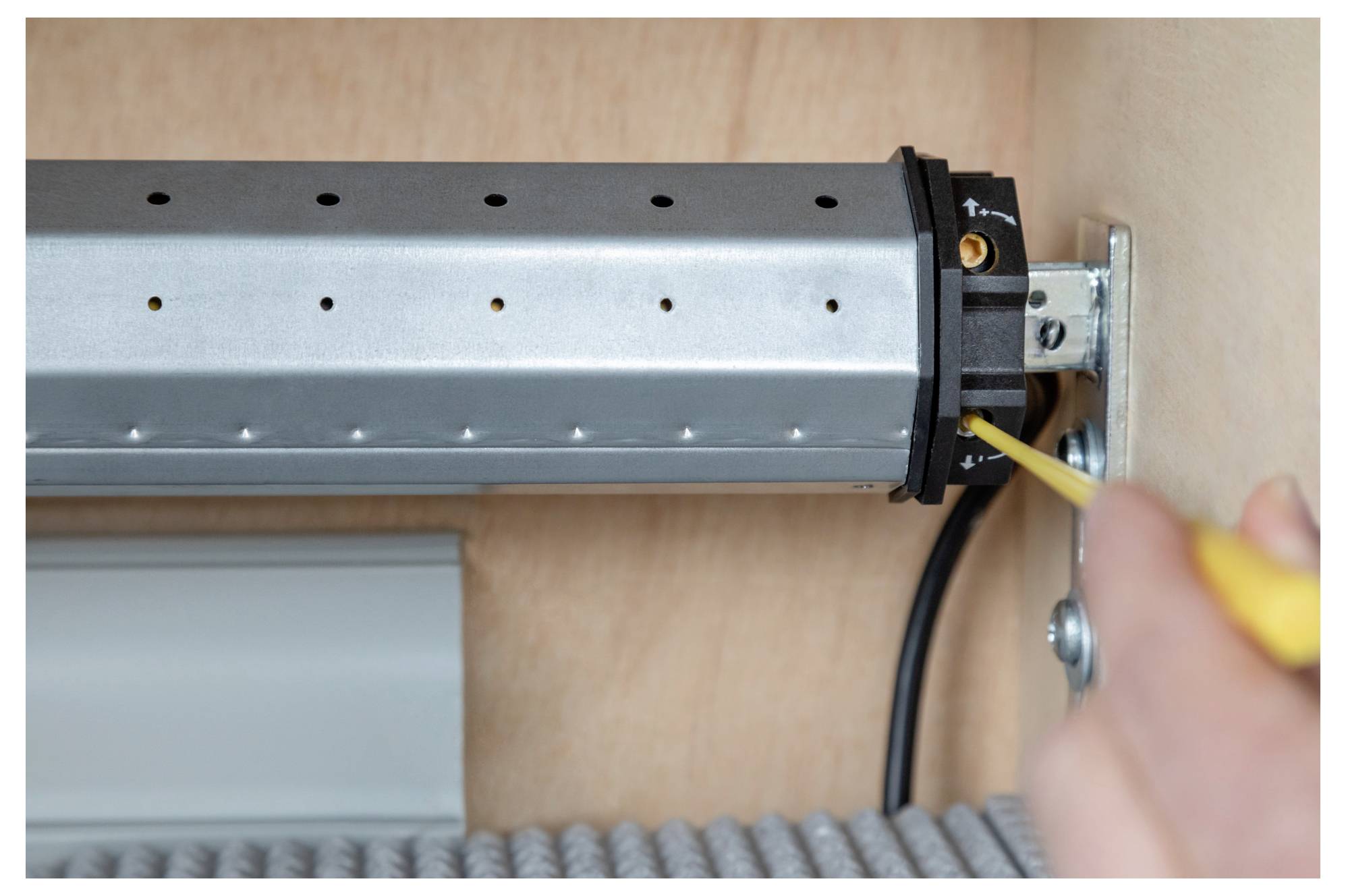 Eine Person zieht eine Schraube an einem metallischen mechanischen Teil, das in einem leichten Holzschrank montiert ist, mit einem leuchtend gelben Schraubenzieher fest.