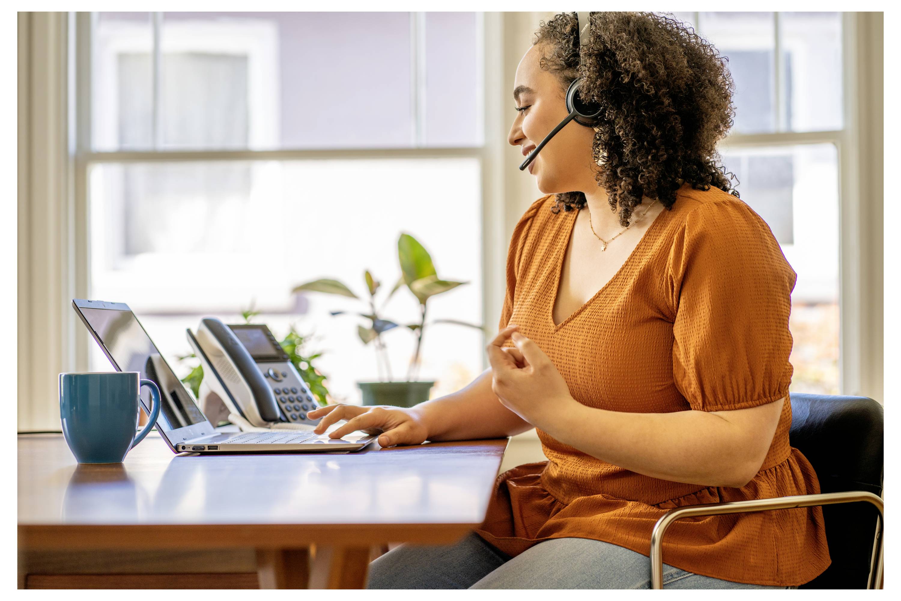 Eine Person, die ein Headset trägt, führt einen Videoanruf auf einem Laptop, sitzt an einem Schreibtisch mit einem Telefon und einer Tasse, vor dem Hintergrund großer Fenster.