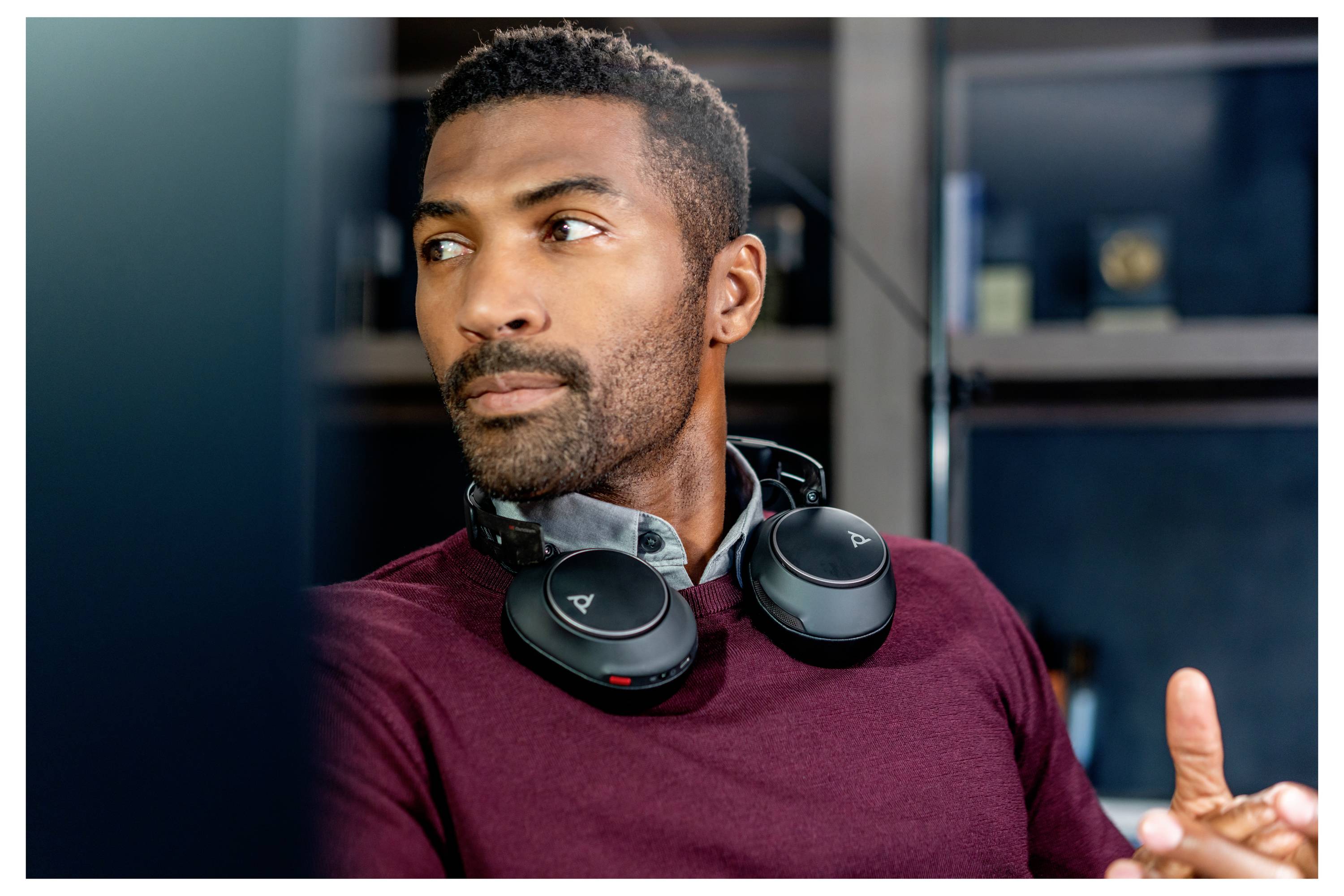Ein Mann mit Kopfhörern um seinen Hals sitzt in einem Büro und blickt nachdenklich zur Seite. Hinter ihm sind Regale mit Büchern sichtbar.