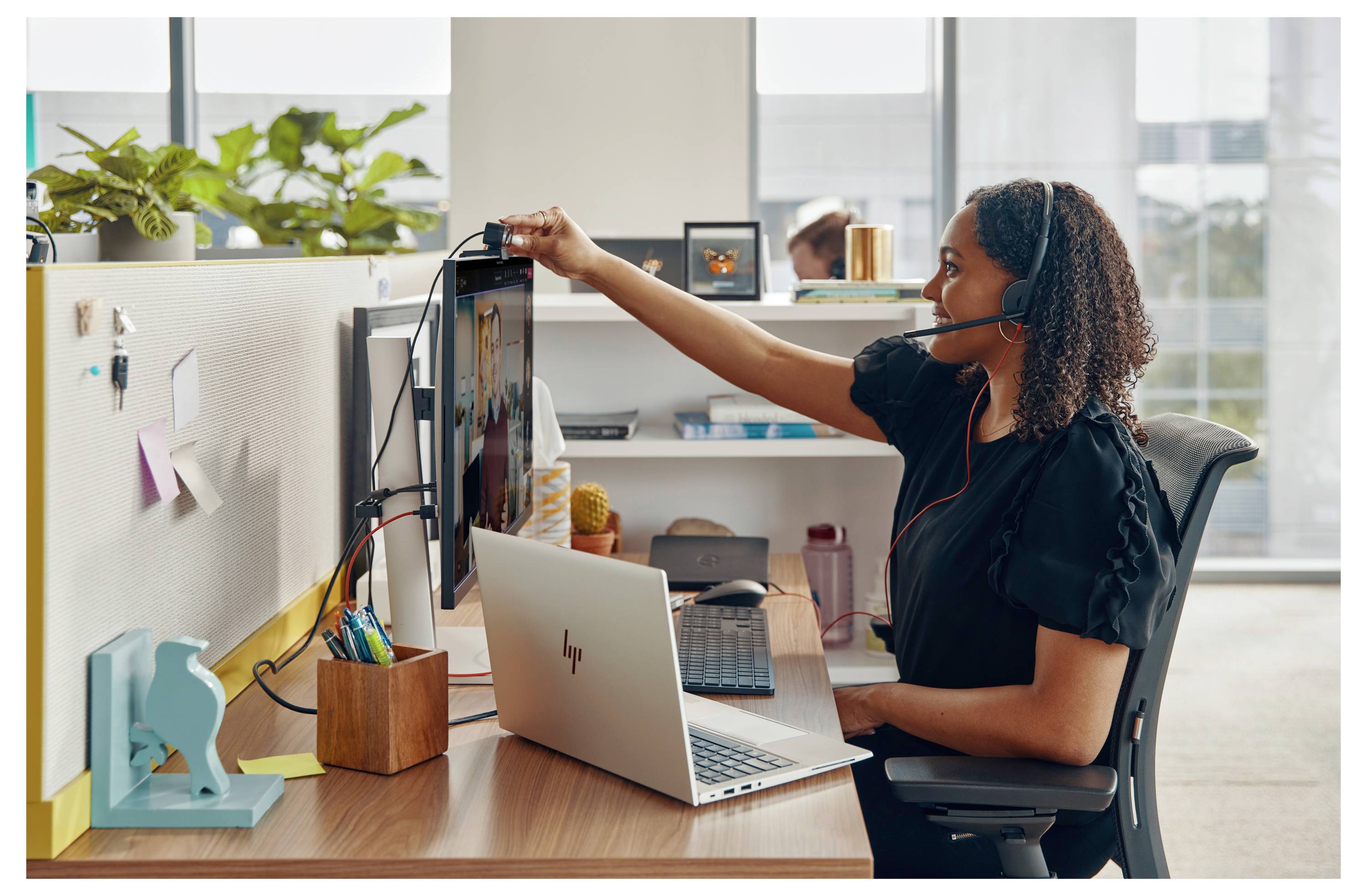 Eine Person mit Kopfhörer arbeitet an einem Laptop an einem Schreibtisch in einem Büro und stellt einen vertikalen Monitor ein. Der Arbeitsbereich ist organisiert und dekoriert.