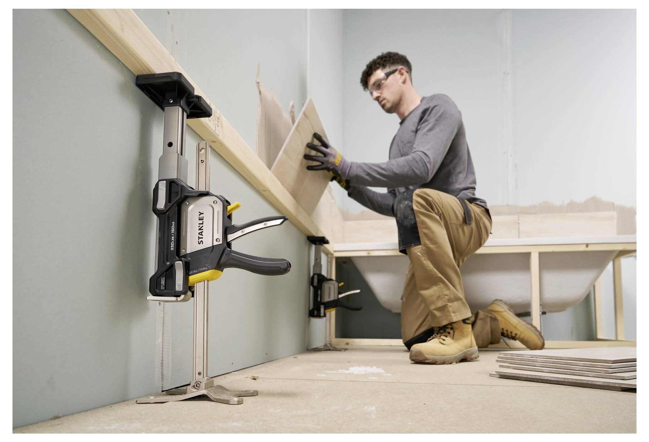 Eine Person kniet auf dem Boden und installiert Wandfliesen in einem Raum. Eine lange Zwinge sichert eine hölzerne Führungsschiene an ihrem Platz und unterstützt die Arbeit.