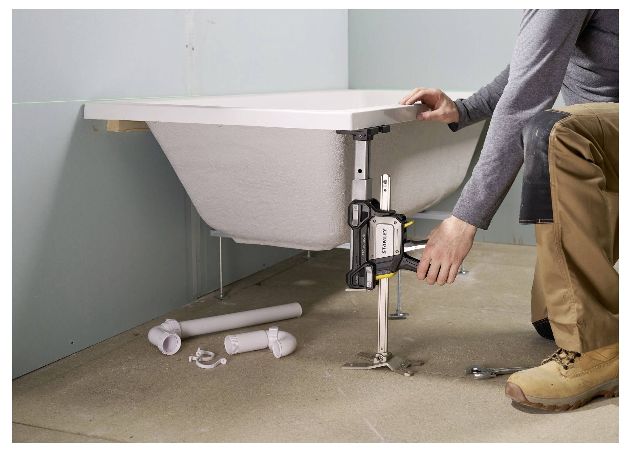 Eine Person installiert eine Badewanne und passt die Höhe mit einem Nivellierwerkzeug an. Rohre für die Sanitärinstallation liegen in der Nähe auf dem Boden.