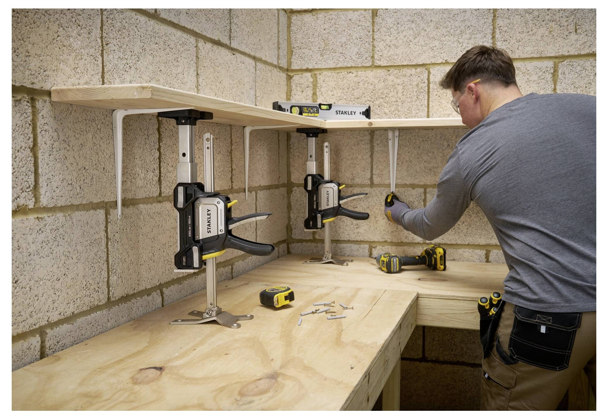 Eine Person benutzt einen Schraubenzieher, um eine Regalbefestigung auf einem Holzregal in einer Werkstatt zu montieren, wobei Werkzeuge und Schraubzwingen auf der Werkbank sichtbar sind.