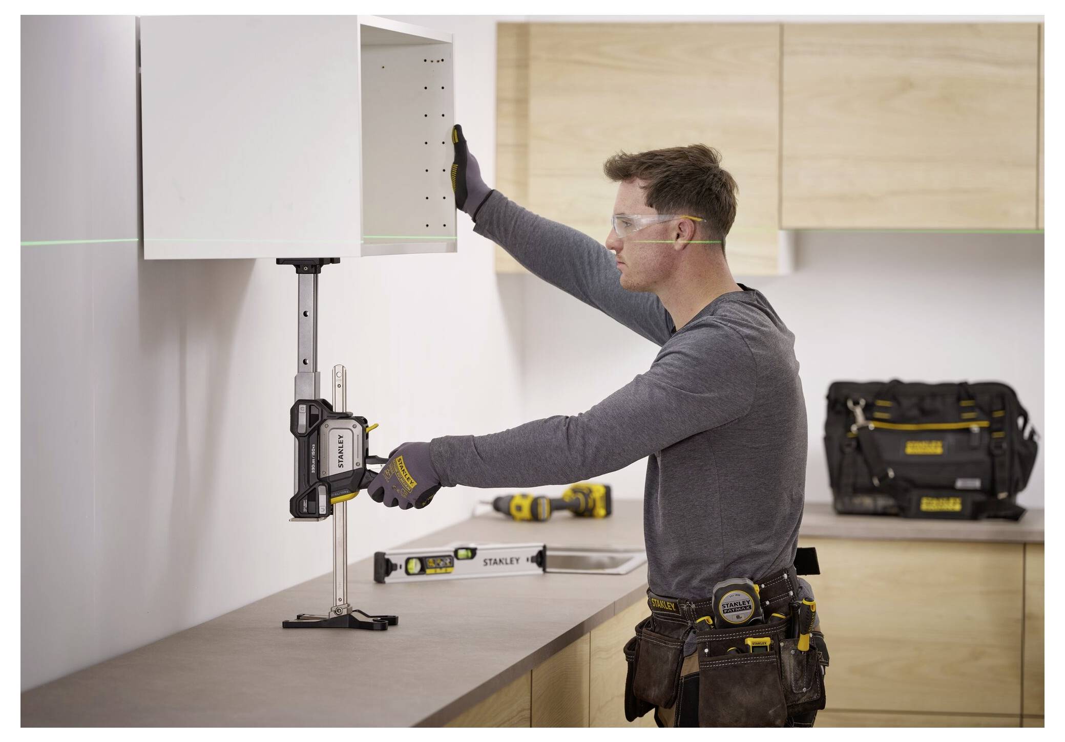 Eine Person, die eine Schutzbrille trägt, verwendet ein Wasserwaagenwerkzeug, um einen Schrank an einer Wand in einer Werkstattumgebung zu installieren. Werkzeuge sind in der Nähe sichtbar.