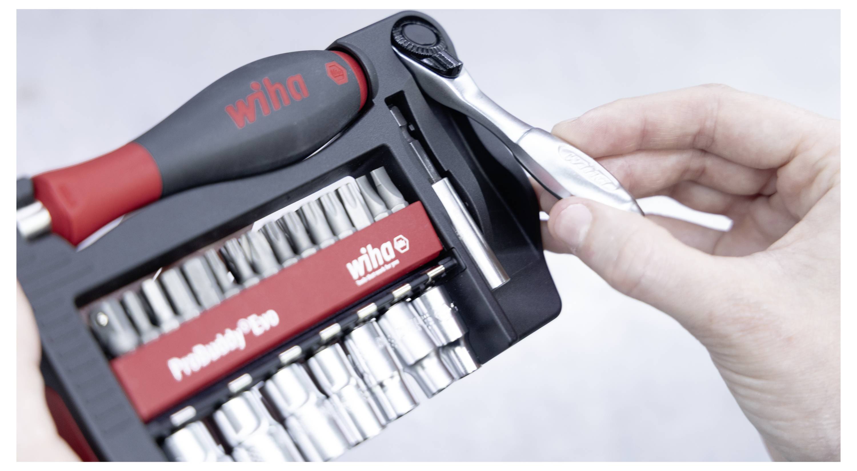 Eine Hand, die einen kleinen Ratschenaufzieher neben einem Präzisionswerkzeugsatz mit verschiedenen Bits und Nüssen hält. Der Werkzeuggriff trägt die Aufschrift „Wiha