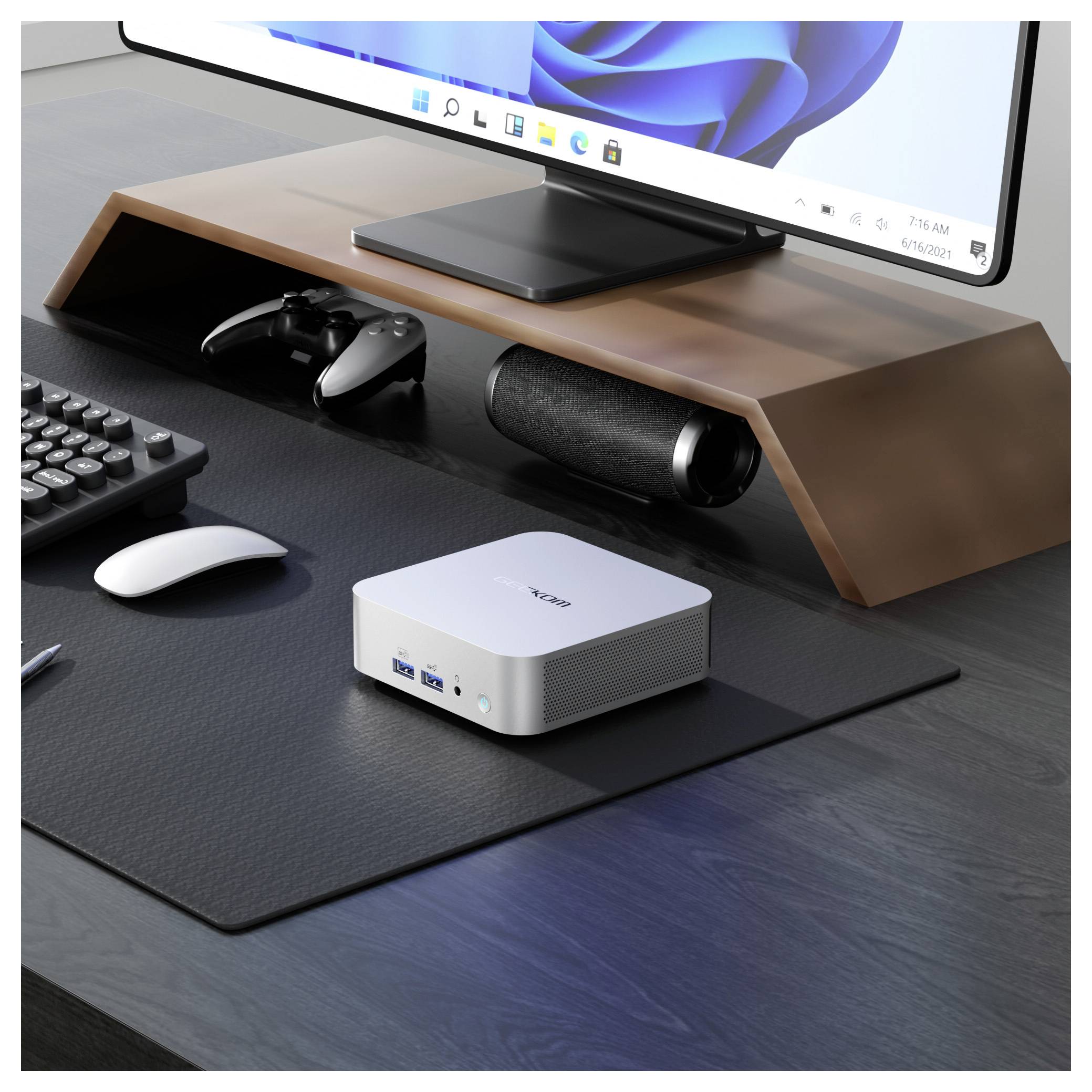 Kompakter weißer Mini-PC auf einem Schreibtisch unter einer Monitorständer, mit Tastatur, Maus, Spielregler und Lautsprecher, was auf ein ordentliches Home-Office-Setup hindeutet.