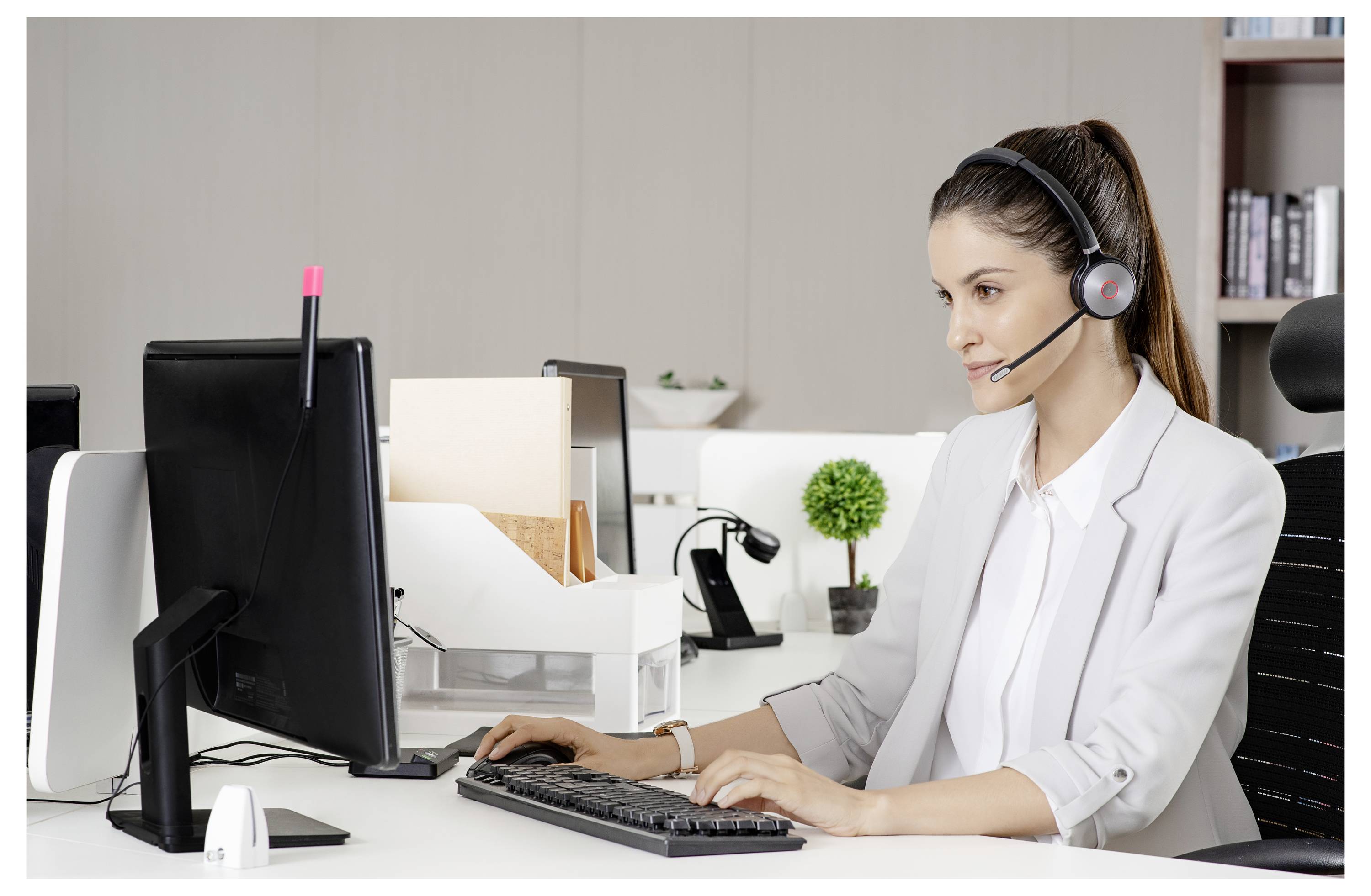 Eine Frau, die ein Headset trägt, arbeitet an einem Computer in einem Büro. Sie ist konzentriert, umgeben von Papieren, einer Pflanze und Büromaterialien.