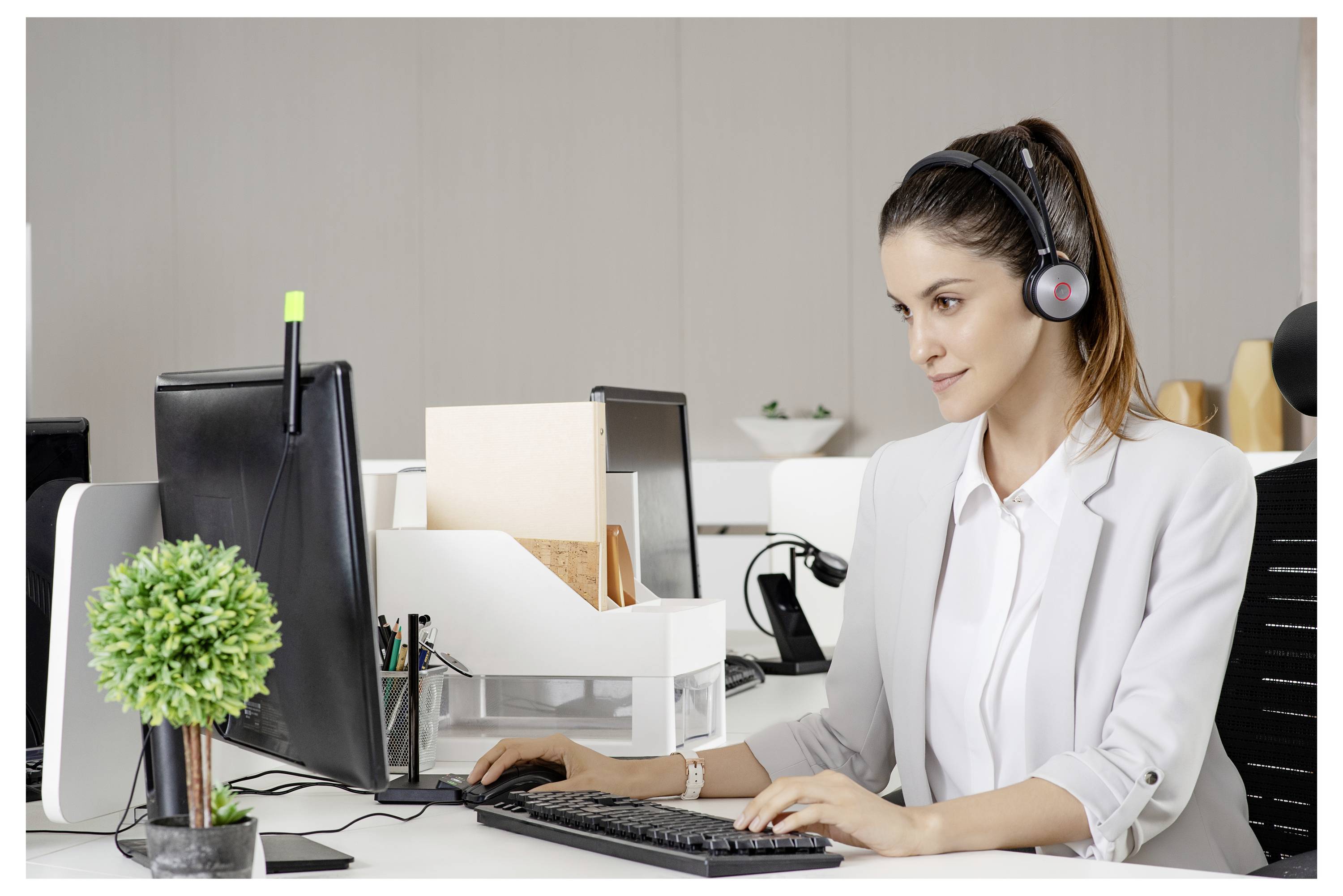 Eine Frau in einem weißen Blazer, die ein Headset trägt, arbeitet an einem Computer in einem modernen Büro, mit einer kleinen Pflanze auf dem Schreibtisch.