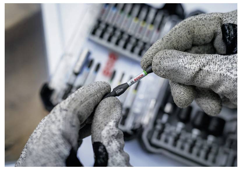 Die behandschuhten Hände einer Person halten einen Bosch-Bohrer, während im unscharfen Hintergrund ein Werkzeugsatz zu sehen ist. Der Fokus liegt auf dem Bohrer und den Handschuhen.