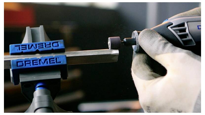 Eine Nahaufnahme einer Person, die ein Schleifwerkzeug verwendet, um die Kante eines in einem blauen Schraubstock eingespannten Metallstabs zu schleifen, wobei die Präzision des Werkzeugs demonstriert wird.