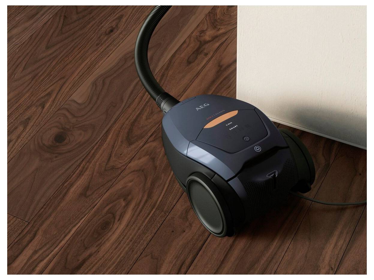 Ein dunkelblauer Staubsauger auf einem Holzboden, positioniert neben einer weißen Wand. Sein Kabel erstreckt sich von der Maschine.