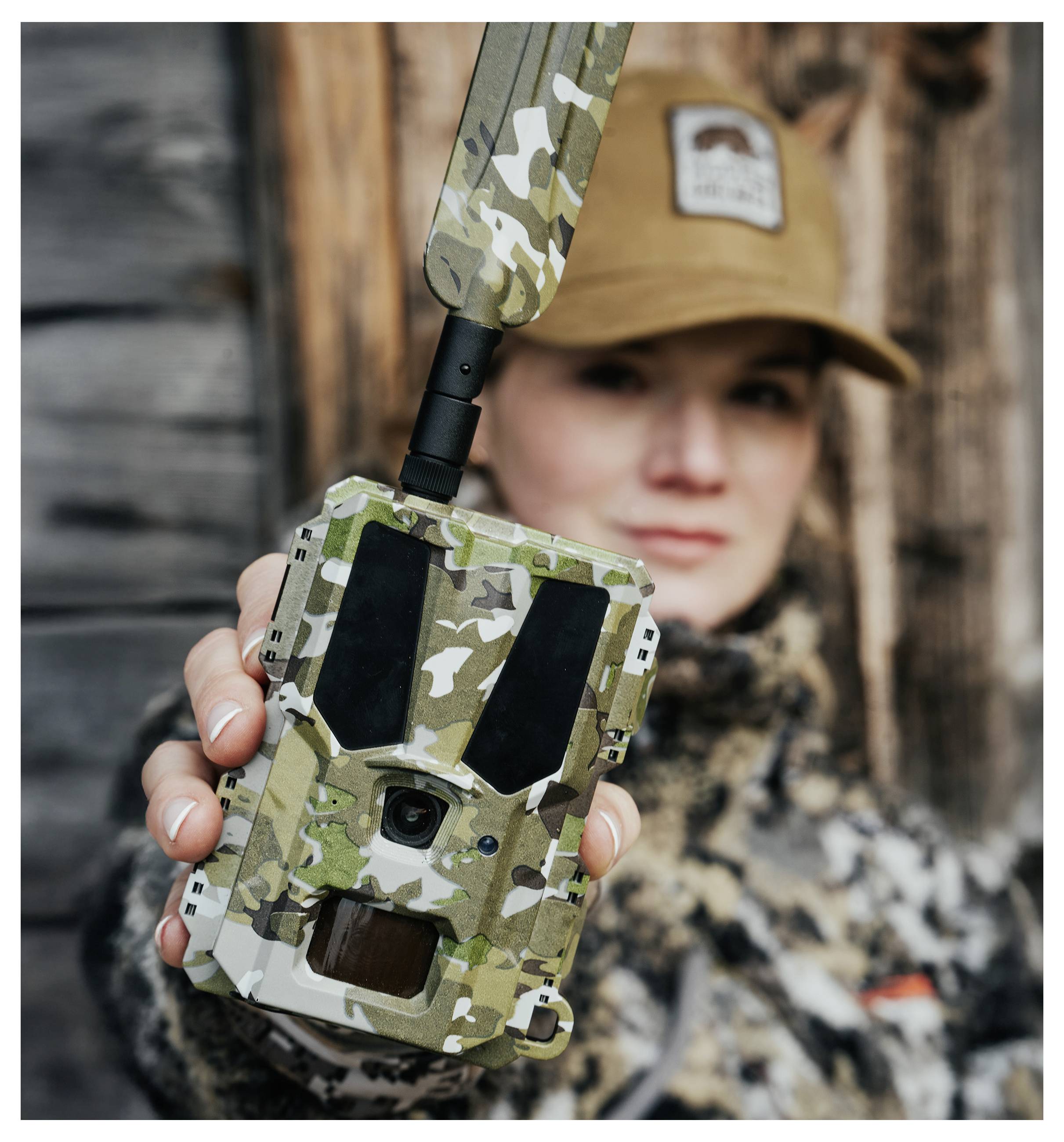 Eine Person in Tarnkleidung hält eine getarnte Wildkamera mit einer Antenne, was auf Außenbeobachtung oder Wildtierüberwachung hindeutet.