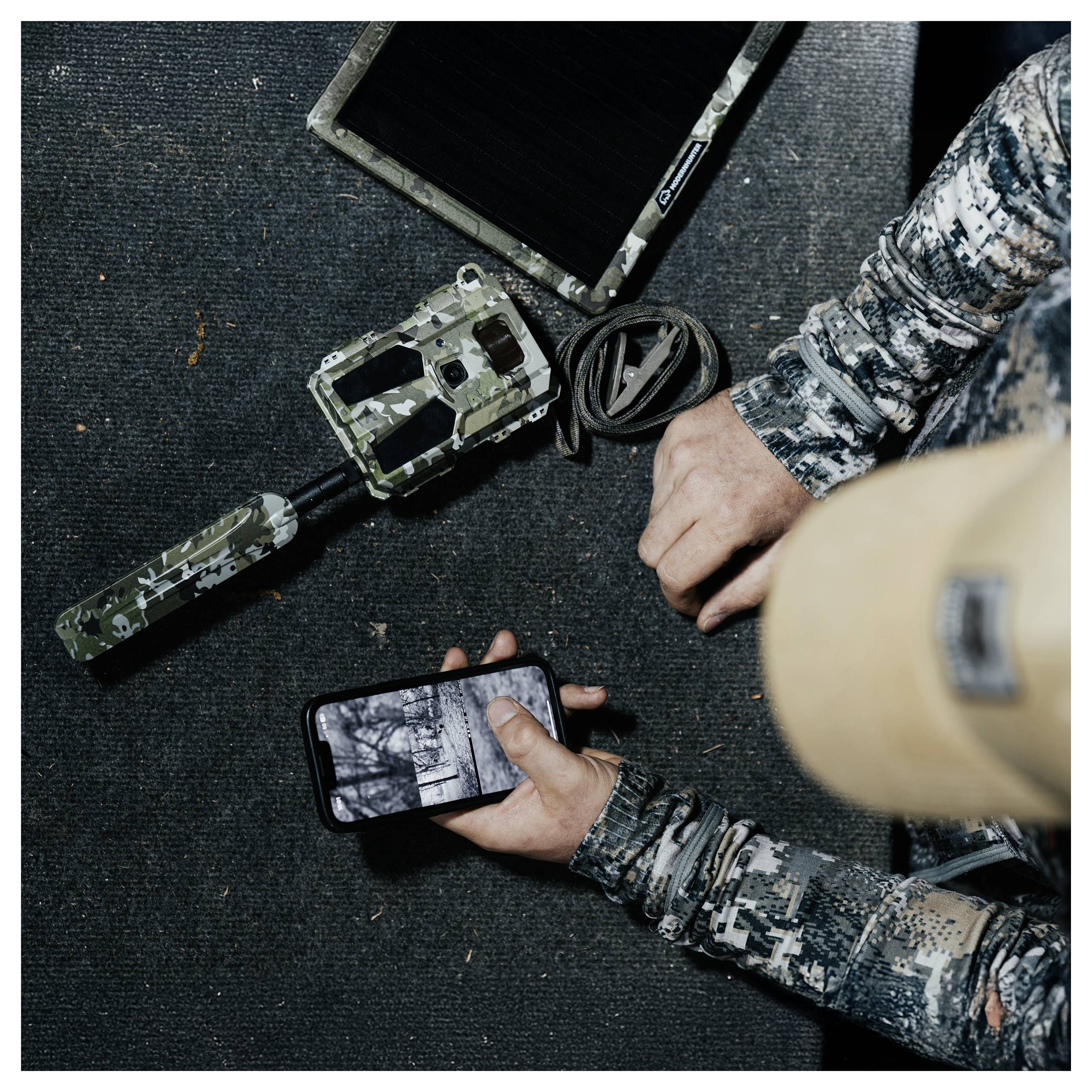 Eine Person in Tarnausrüstung überprüft ein Wildkamera-Bild auf einem Smartphone, während ähnliche getarnte Ausrüstung in der Nähe liegt.