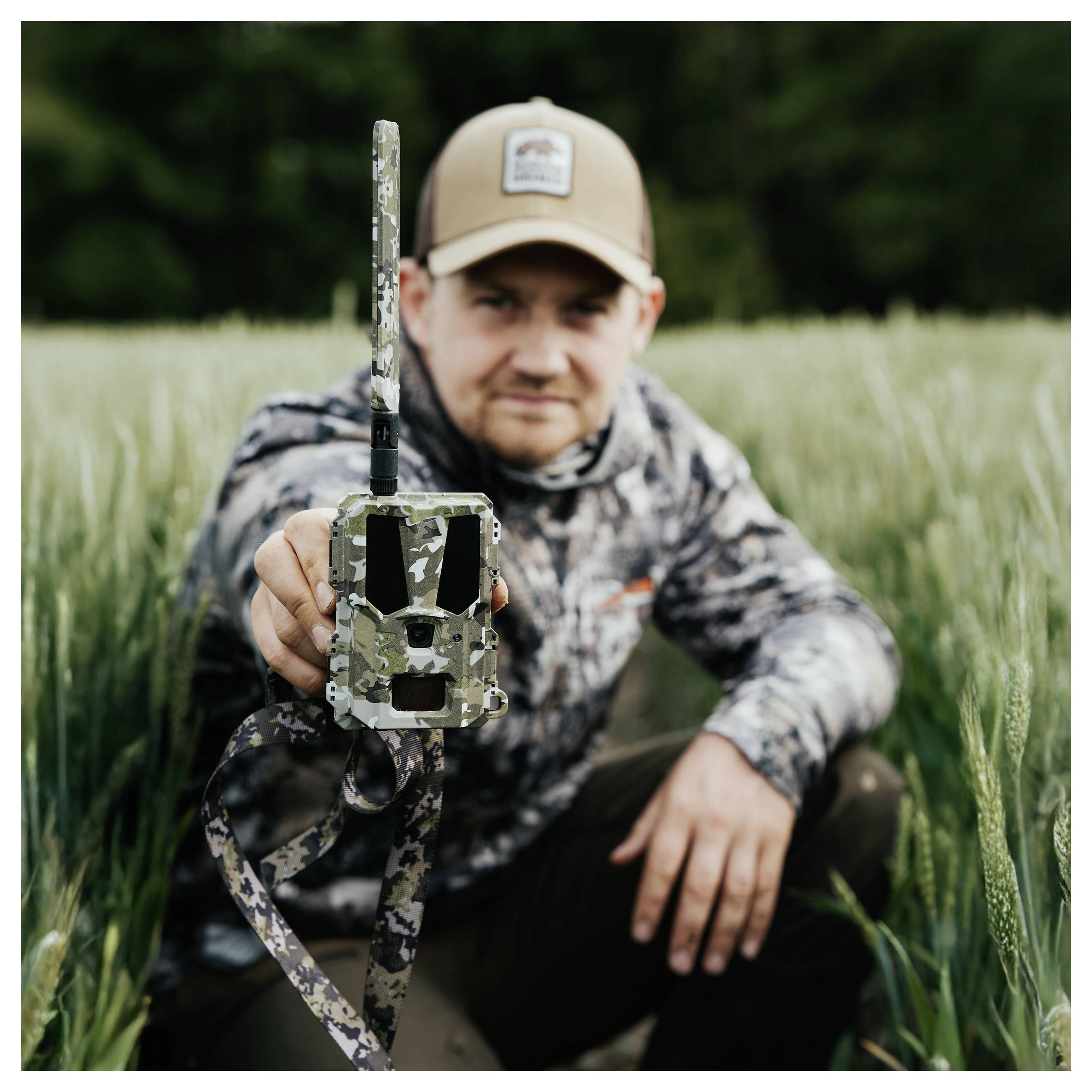 Eine Person in Tarnkleidung und mit einer Kappe kniet in einem Feld und hält eine Tarn-Wildkamera in Richtung des Betrachters.