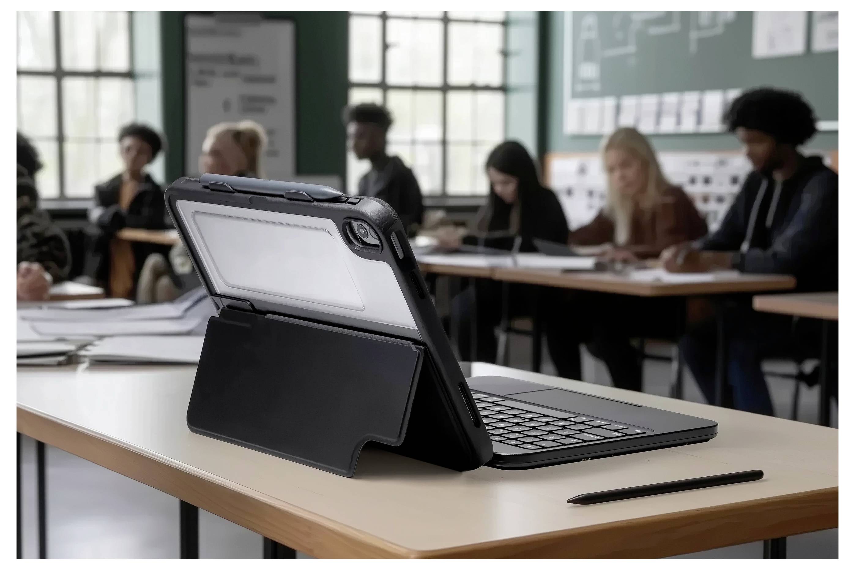 Ein digitales Tablet mit Tastatur liegt auf einem Schreibtisch in einem Klassenzimmer. Schüler sitzen im Hintergrund und beschäftigen sich mit Lernaktivitäten.