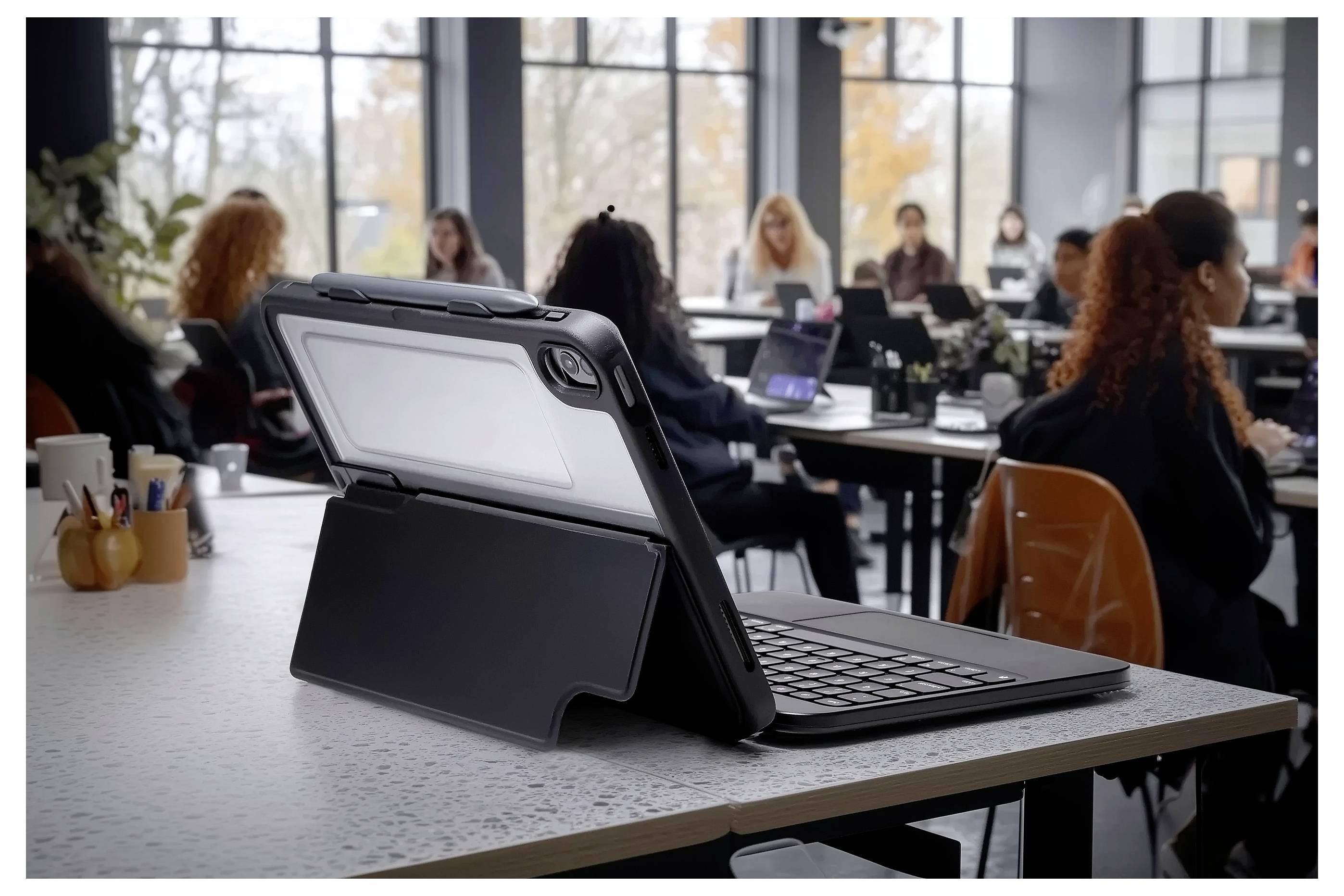 Ein Tablet mit Tastaturcase liegt im Vordergrund auf einem Schreibtisch, während Schüler im Hintergrund an Schreibtischen in einer Klassenzimmeratmosphäre sitzen.