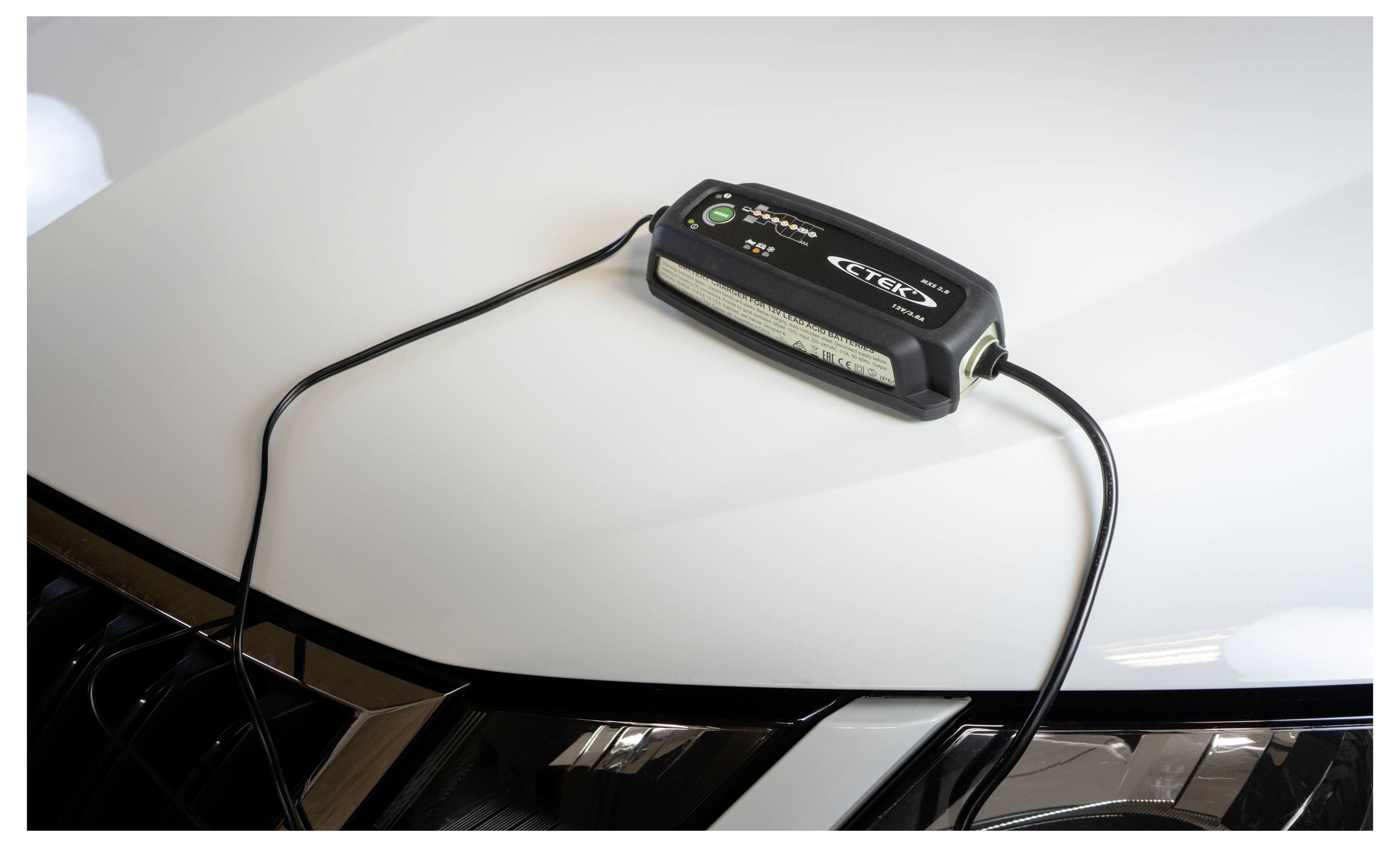 Ein schwarzes Batterieladegerät sitzt auf der Motorhaube eines weißen Fahrzeugs mit angeschlossenen Kabeln, was darauf hindeutet, dass es die Batterie des Fahrzeugs auflädt.