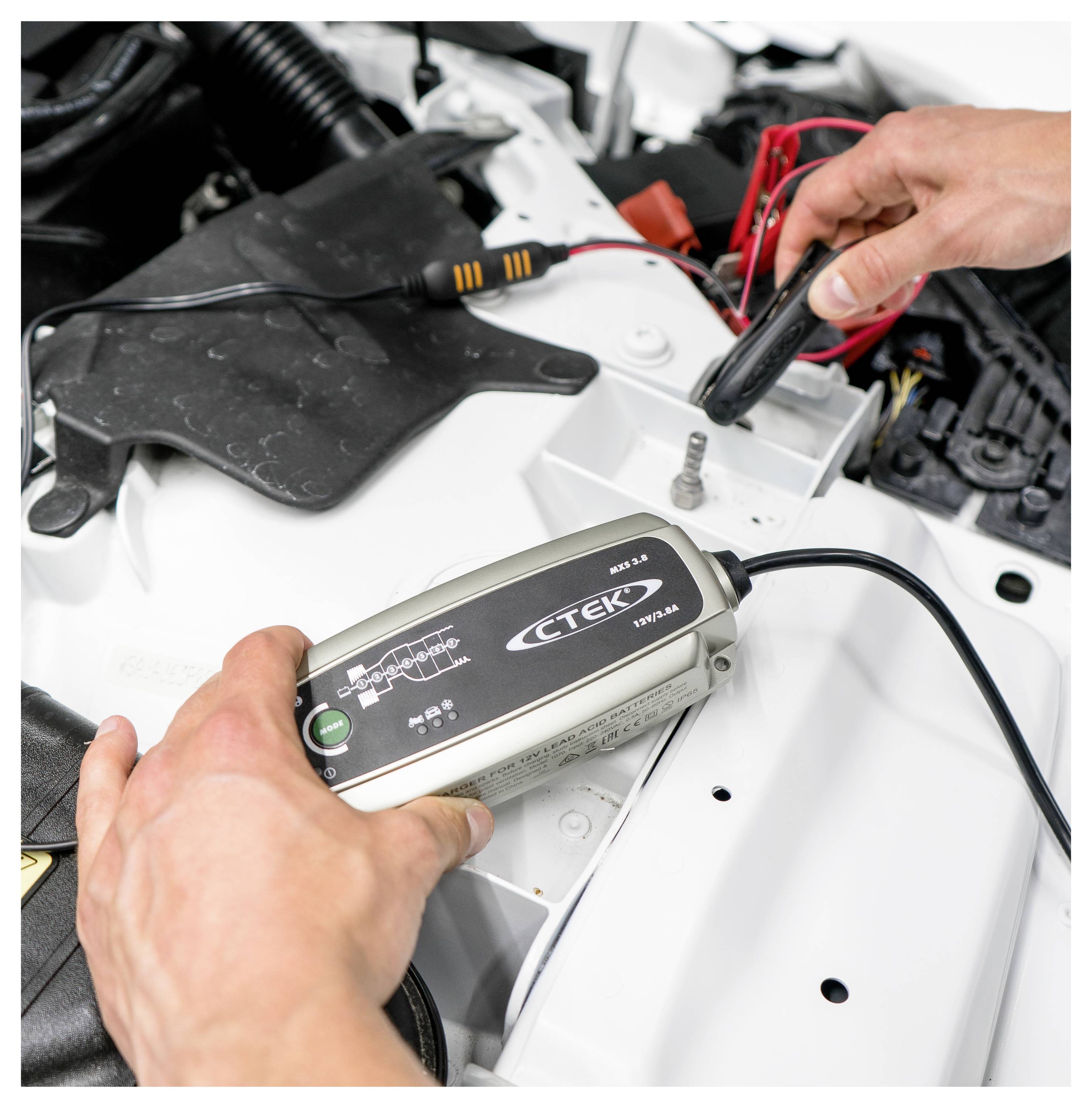 Eine Person verbindet ein CTEK-Ladegerät mit einer Autobatterie, wobei rote und schwarze Kabel sichtbar sind. Die Motorhaube ist geöffnet.