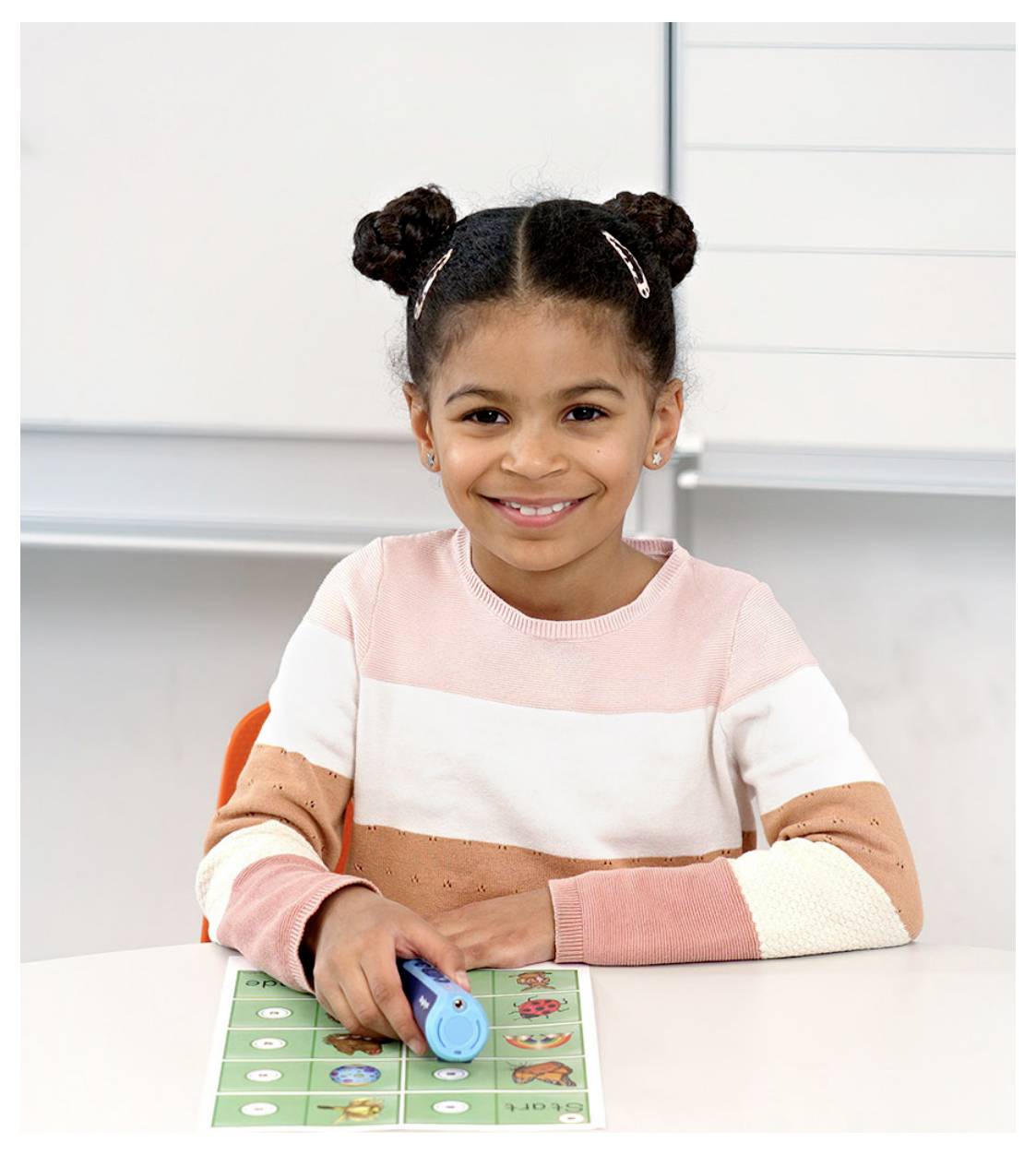 Ein lächelndes Kind mit geflochtenen Haaren sitzt an einem Schreibtisch und hält einen blauen stiftartigen Gegenstand über einem bunten Lernbrett.