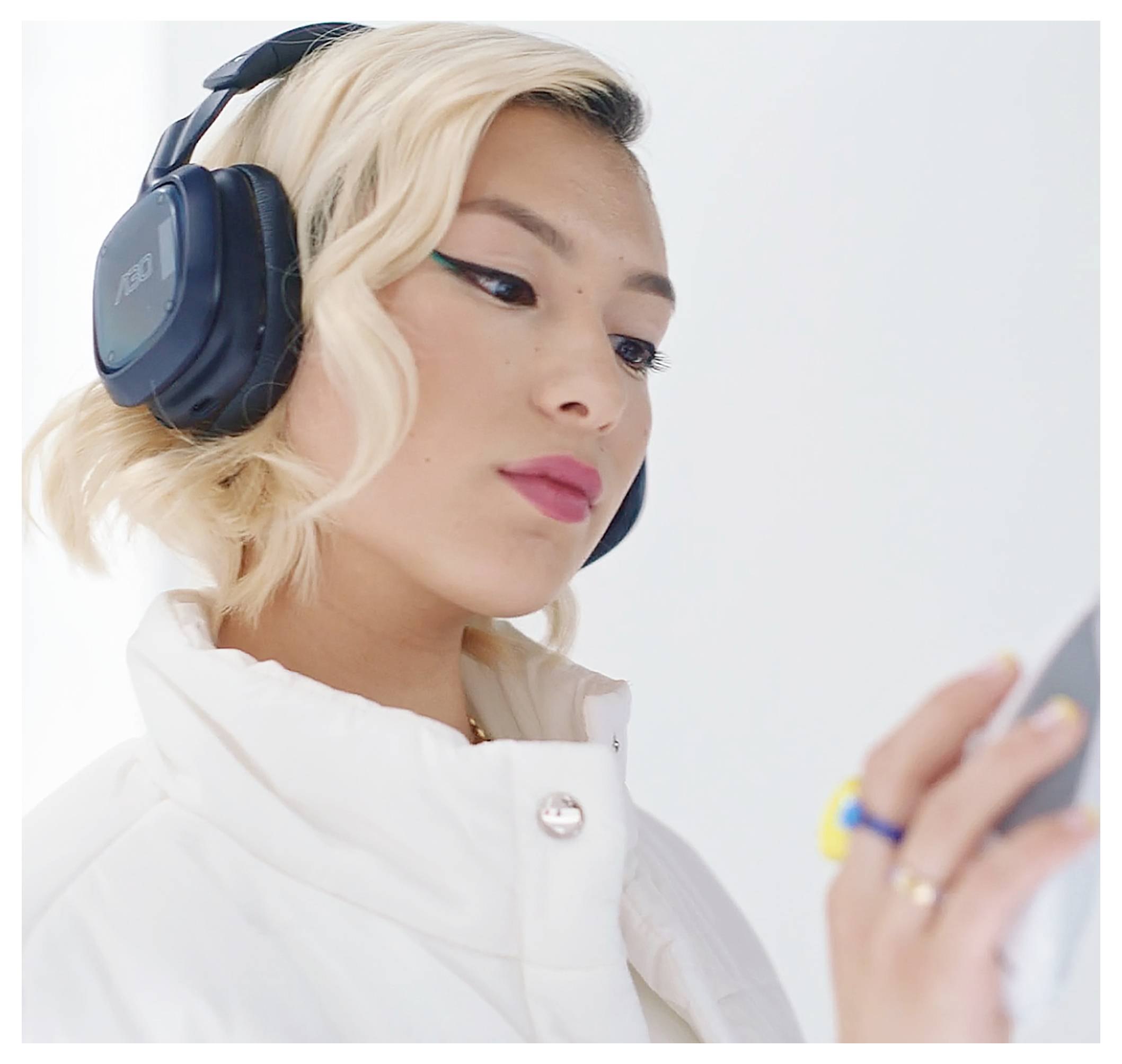 Nahaufnahme einer blonden Frau mit großen Kopfhörern und einer weißen Jacke, die auf ihr Smartphone schaut und es gegen einen hellen, einfachen Hintergrund benutzt.