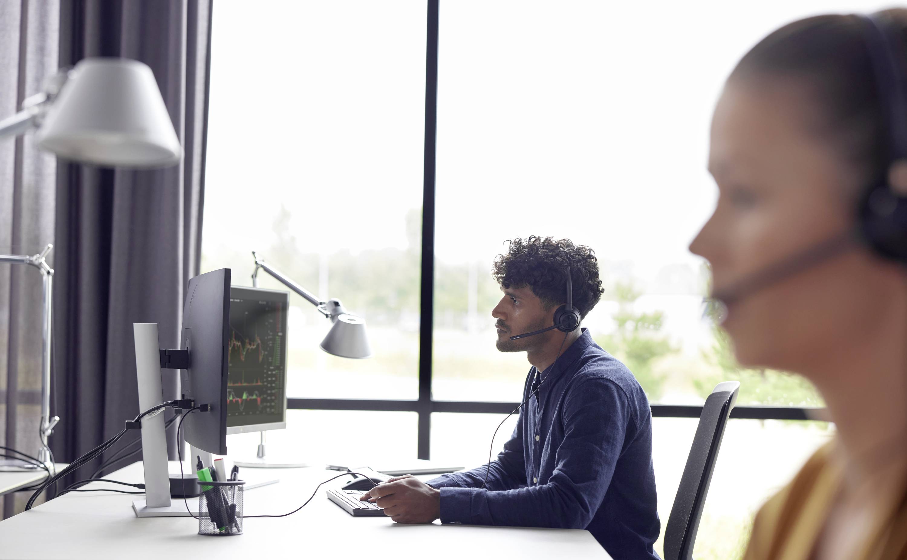 Zwei Kopfhörer tragende Büroangestellte an Schreibtischen, eine konzentriert auf einen Computerbildschirm mit Daten in einem hellen modernen Arbeitsplatz.