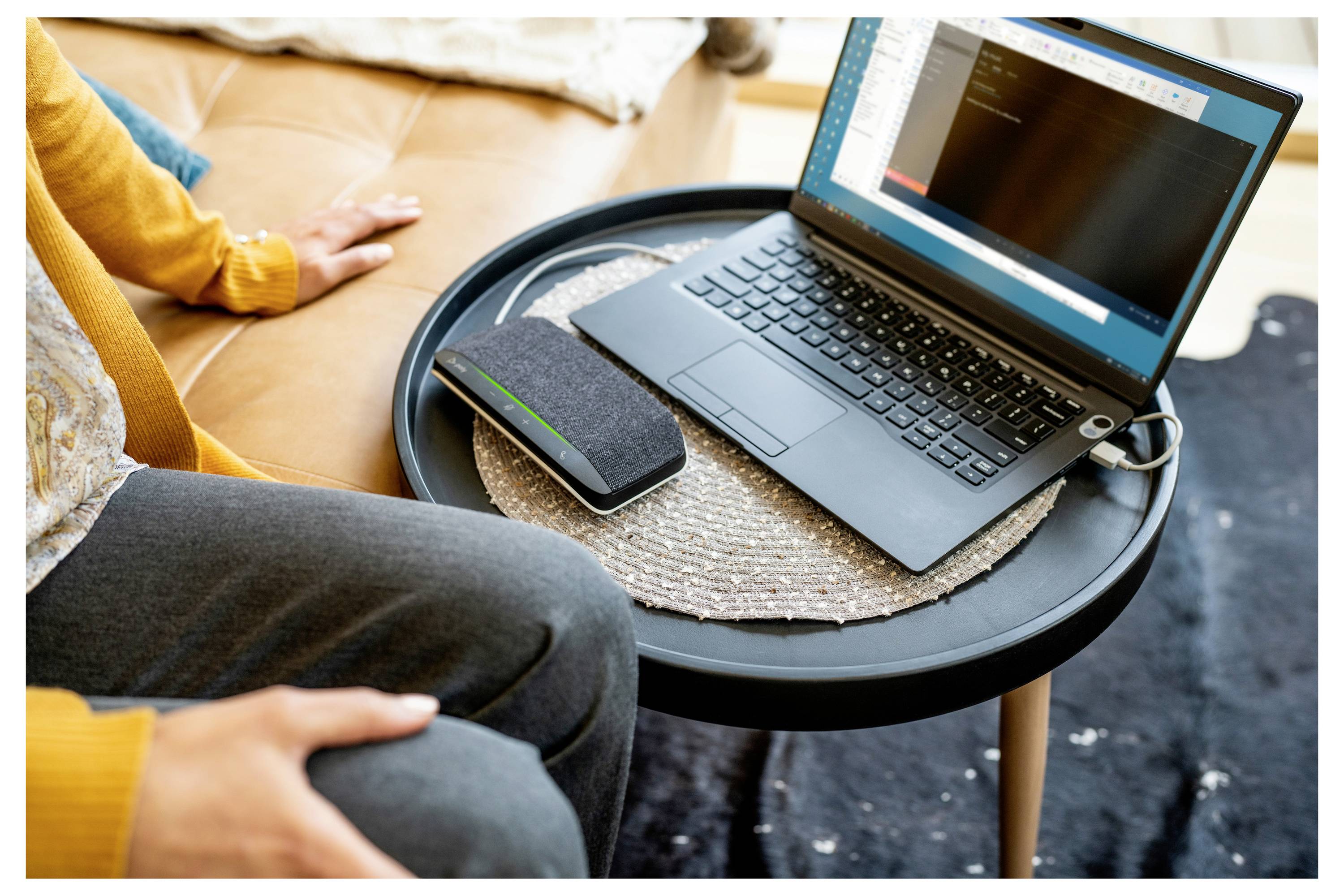 Eine Person sitzt neben einem aufgeklappten Laptop auf einem kleinen Tisch, mit einem Smartphone und Notizbuch in der Nähe in einer häuslichen Umgebung.