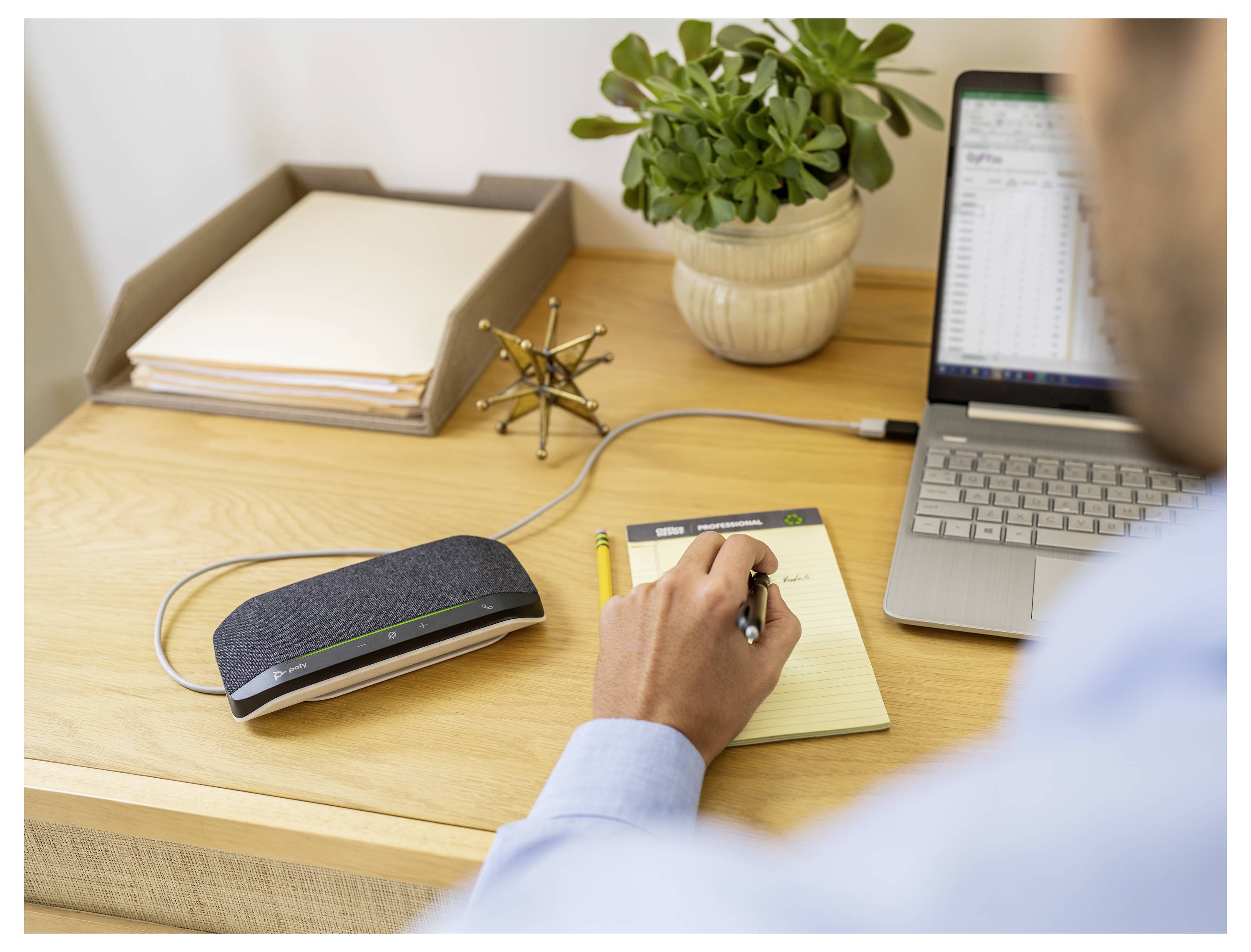 Eine Person, die an einem Schreibtisch neben einem Laptop, Papieren, einer Pflanze und einem verbundenen Lautsprechergerät Notizen schreibt.
