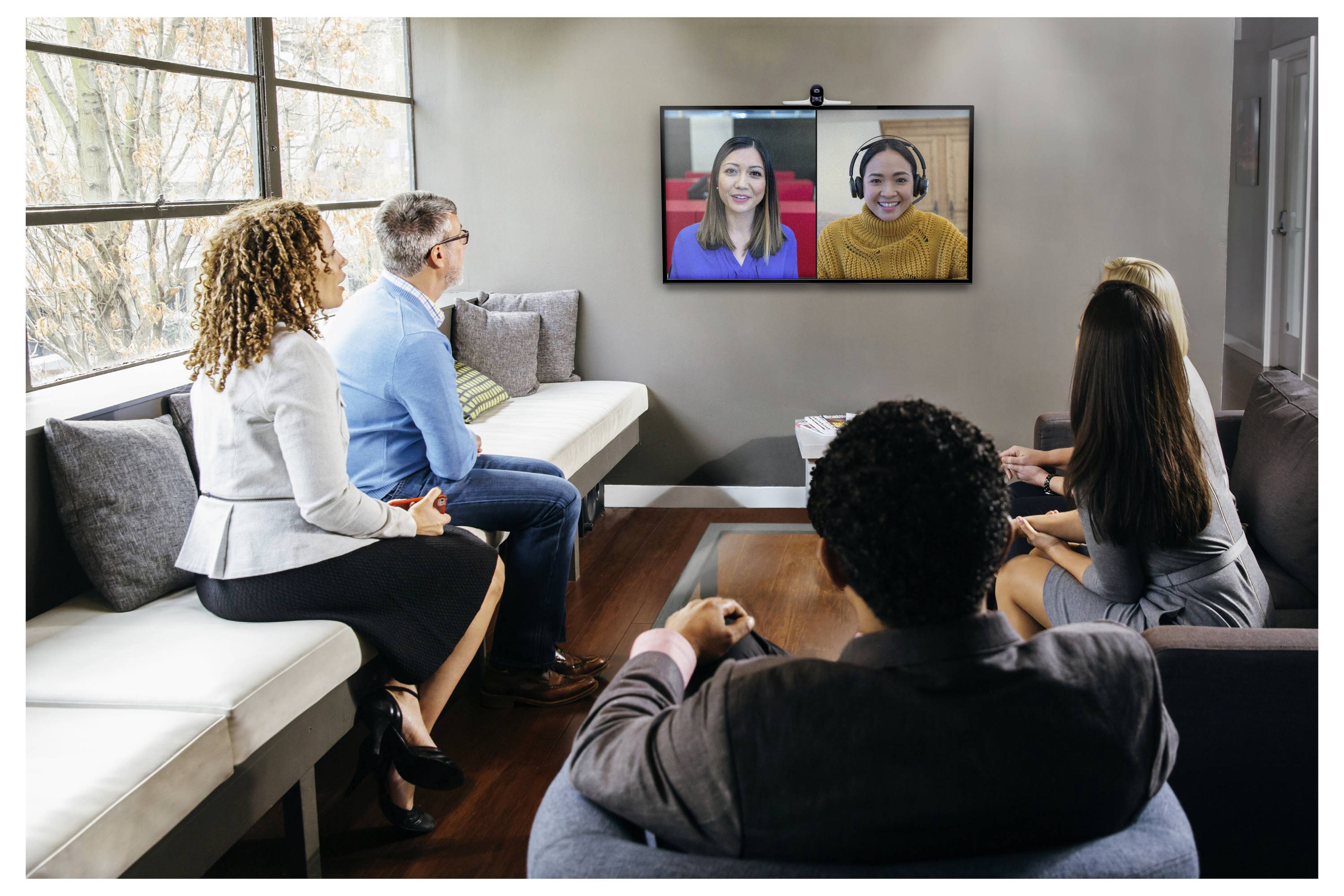 Gruppenvideokonferenz mit vier Personen, die in einem Wohnzimmer sitzen und zwei externe Teilnehmer auf einem an der Wand montierten Fernseher beobachten.