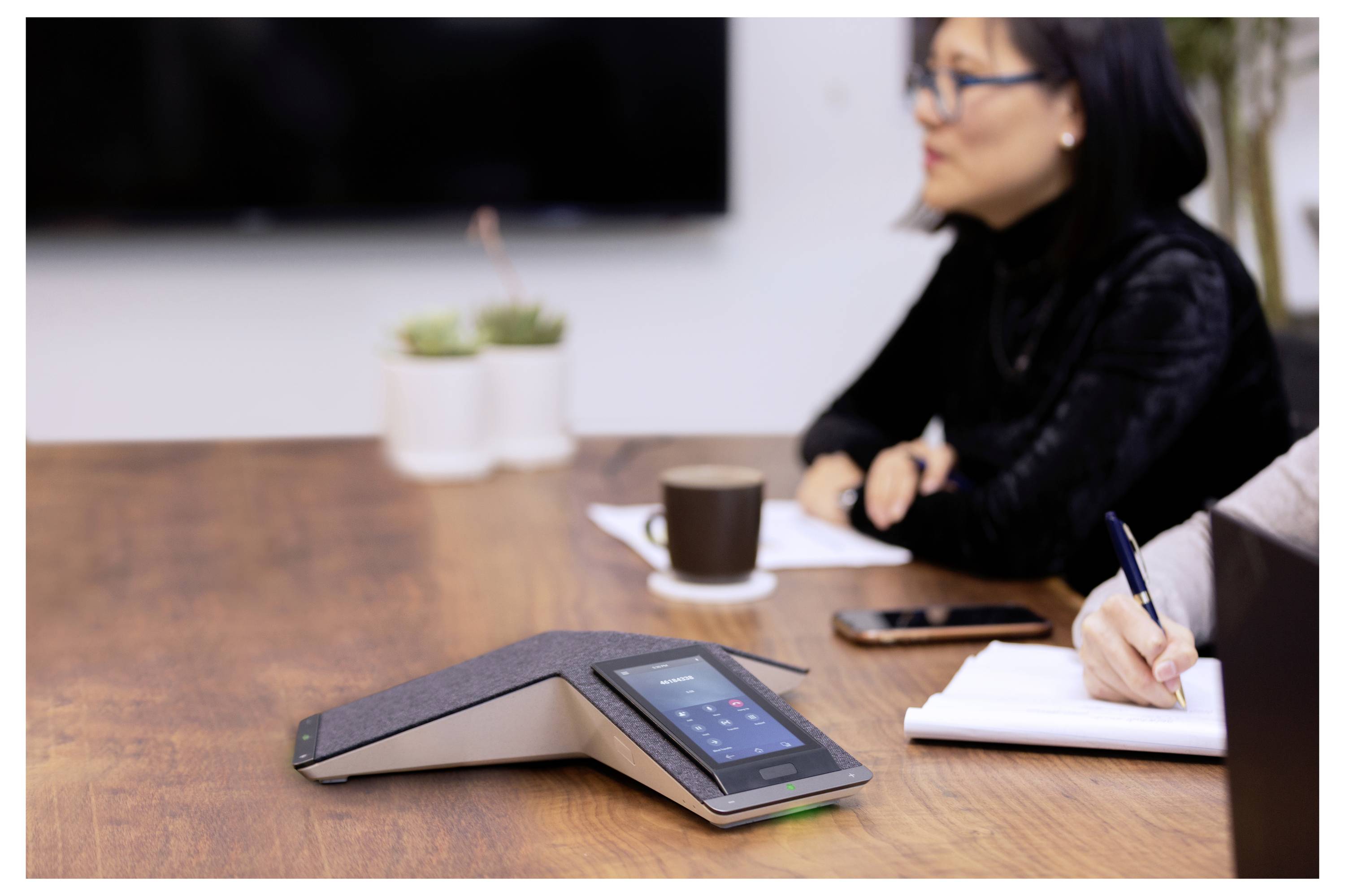 Konferenzlautsprecher auf einem Besprechungstisch, mit zwei Personen, die sich Notizen machen, und einer Kaffeetasse im Hintergrund