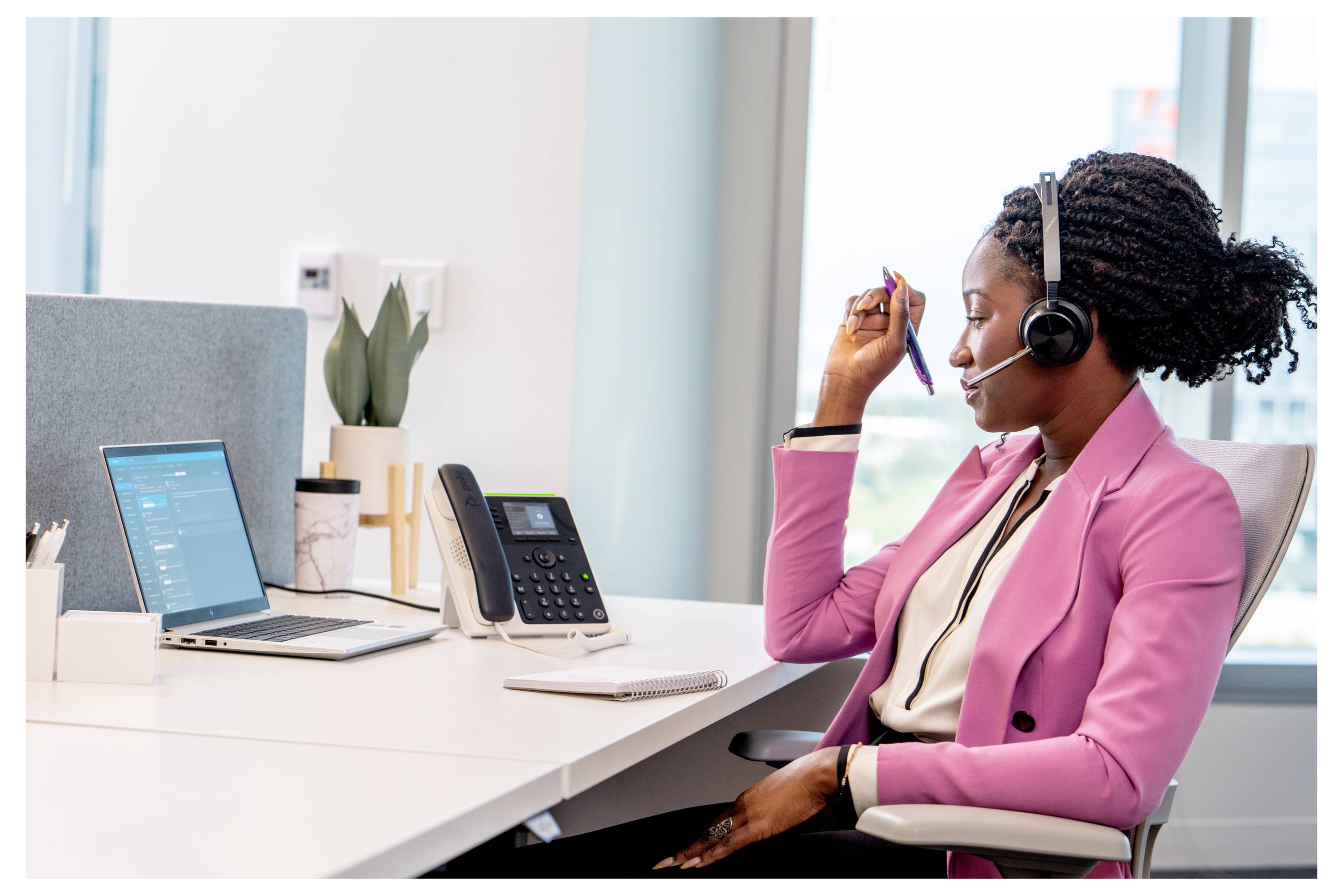 Eine Frau mit Headset sitzt an einem Schreibtisch in einem Büro und spricht in ein Mikrofon, während sie an einem Laptop und einem Telefon arbeitet, was auf einen Kundendienstanruf hindeutet.