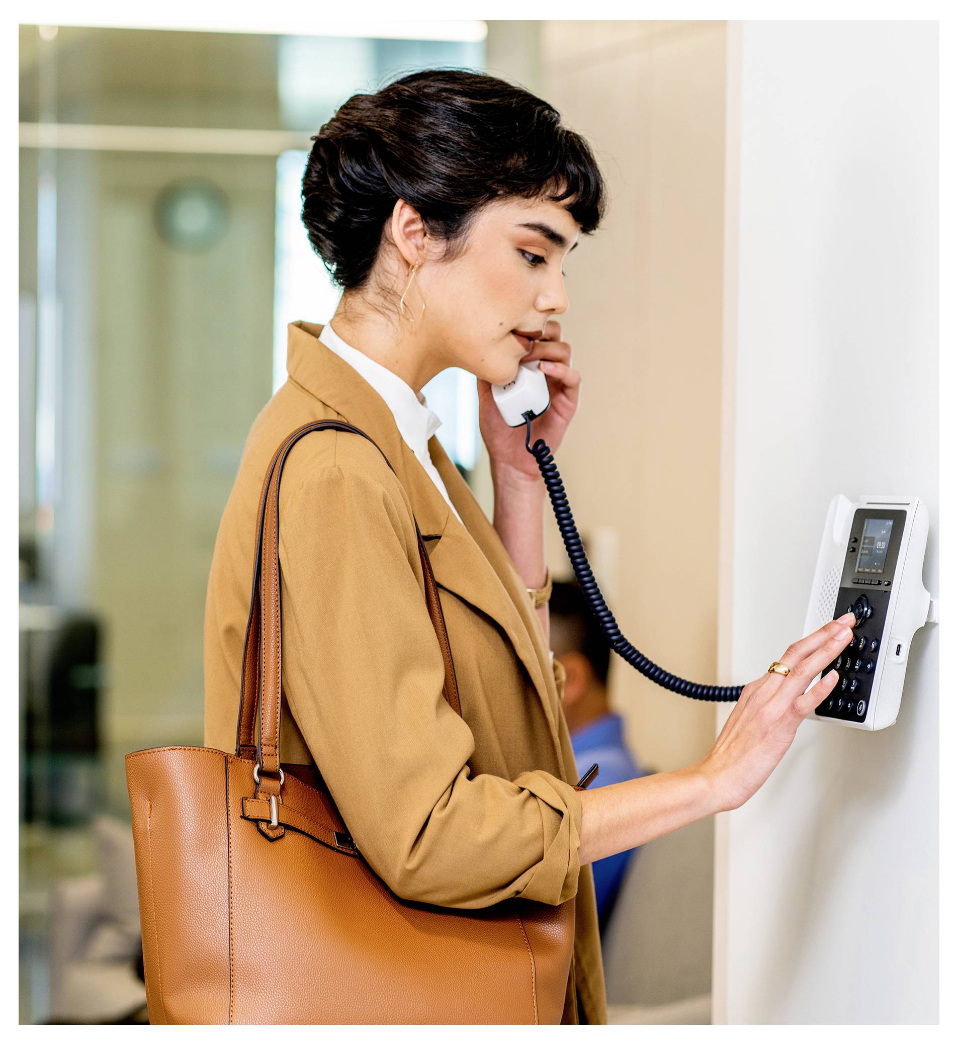 Frau, die ein an der Wand montiertes Bürotelefon benutzt, während sie spricht und eine beige Tragetasche trägt.