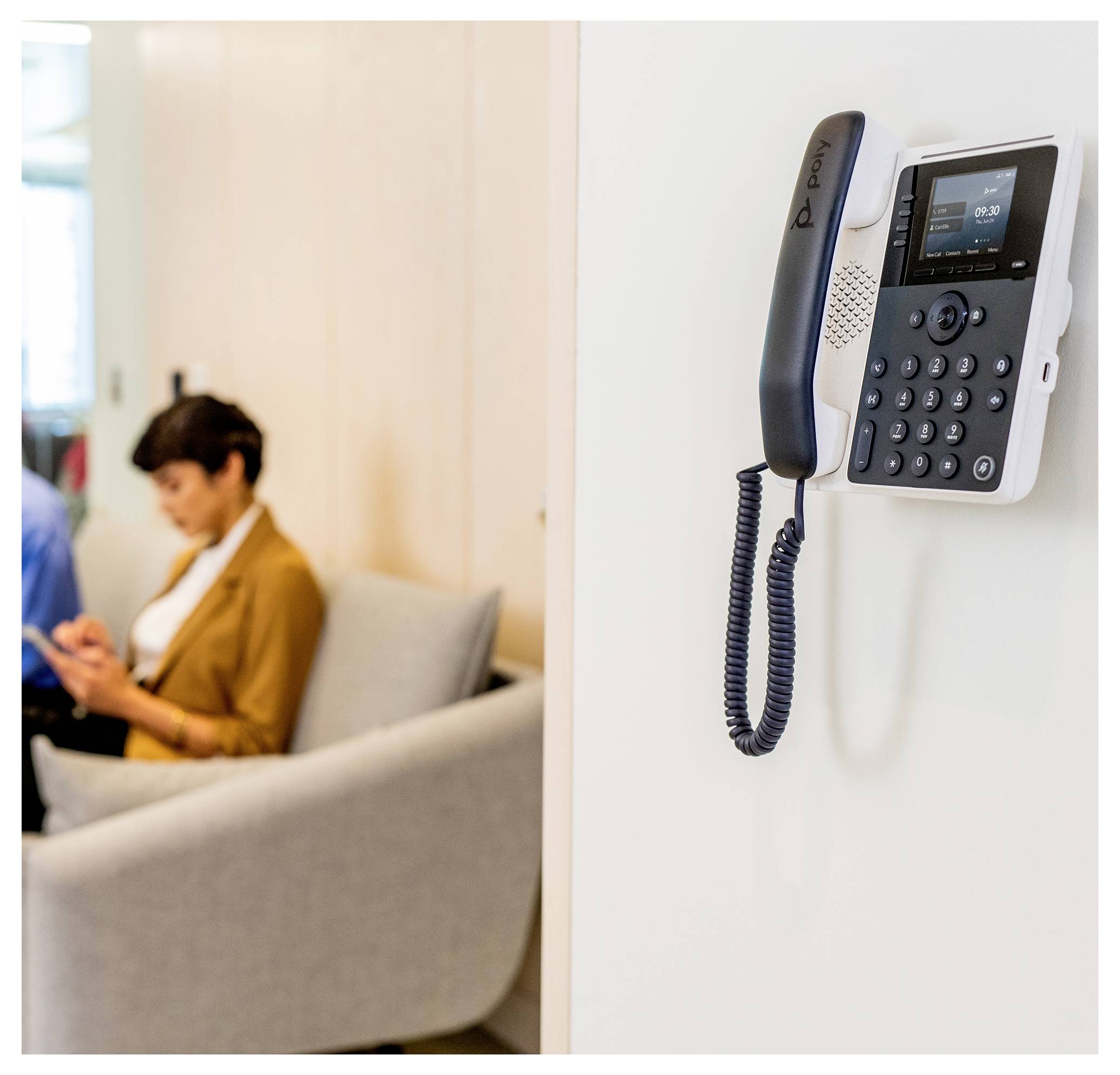 Wandmontiertes Bürotelefon im Vordergrund; verschwommene Person sitzt im Hintergrund auf einem Sofa und benutzt ein Telefon.