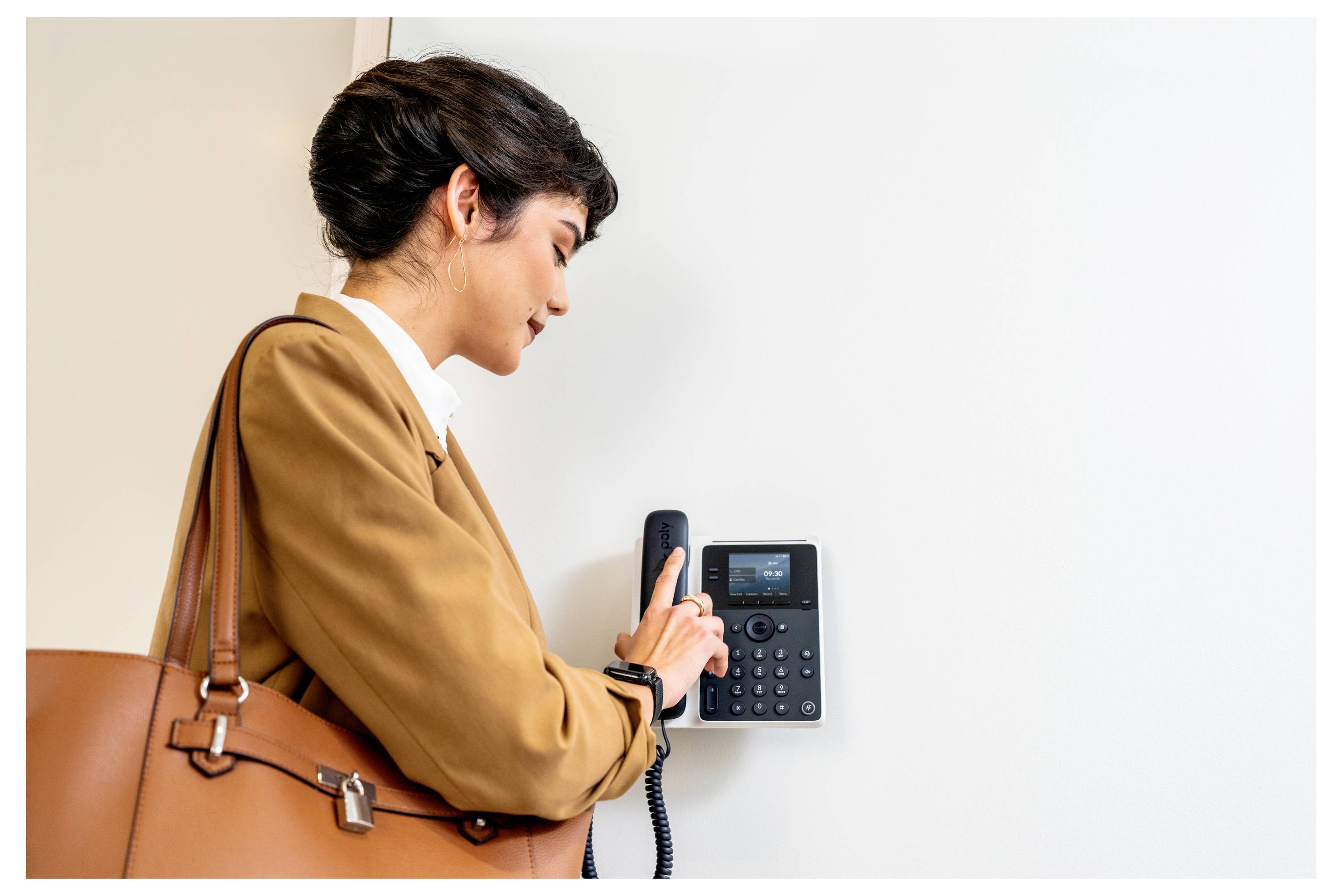 Frau in einem beigen Mantel benutzt ein wandmontiertes Bürotelefon oder eine Gegensprechanlage und hält den Hörer neben einer leeren weißen Wand.
