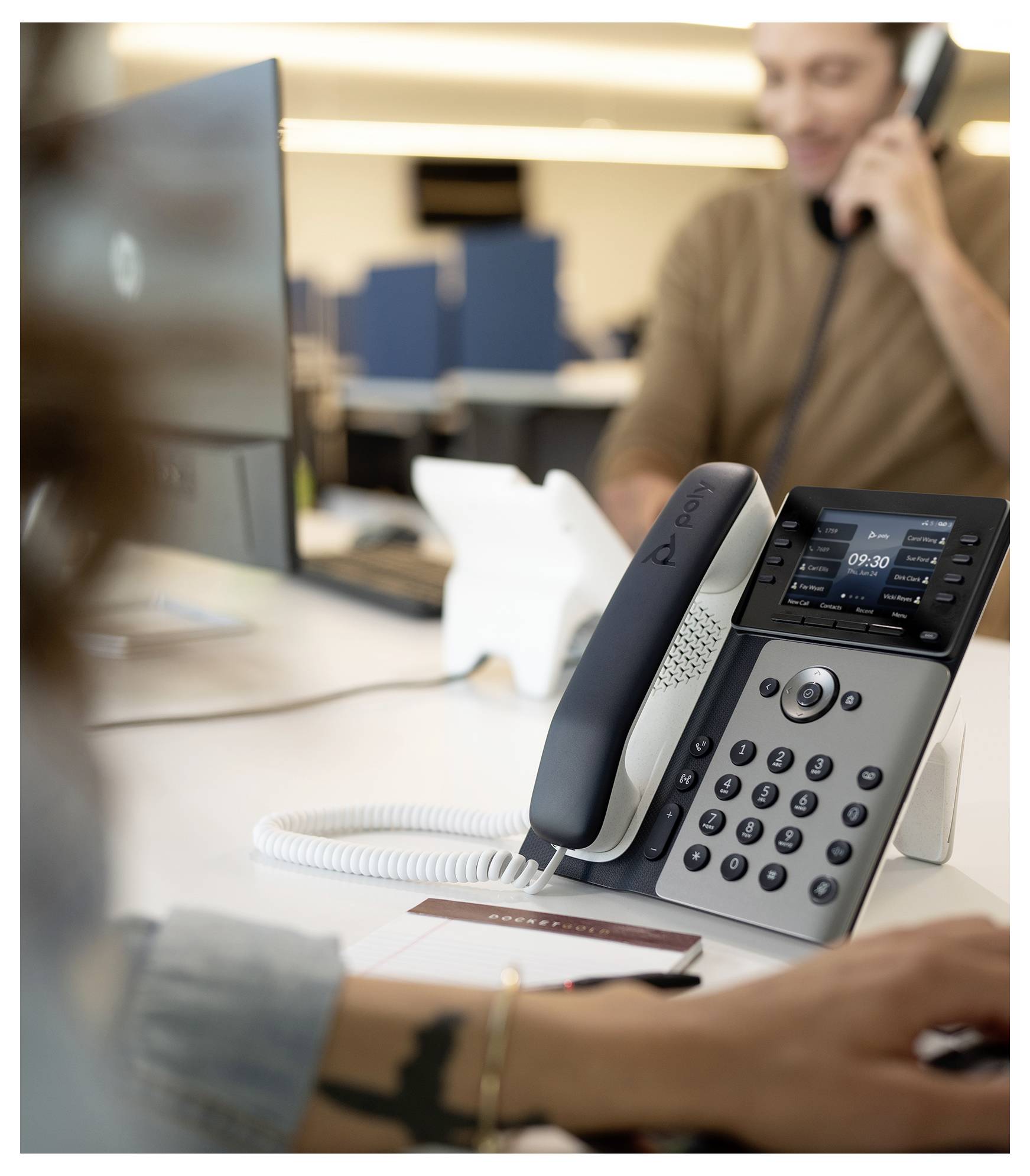 Schreibtischtelefon im Büro im Vordergrund, mit einem unscharfen Mitarbeiter in einem Telefonat und Computermonitor im Hintergrund.